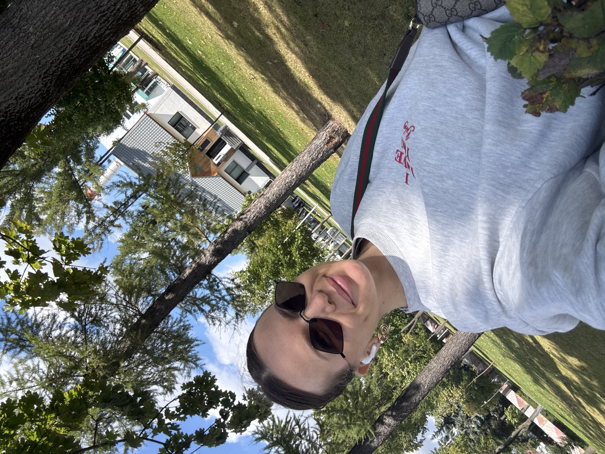 In an outdoor selfie, a woman with dark hair pulled back, wearing sunglasses and white wireless earbuds, smiles at the camera. She is dressed in a grey sweatshirt with a red graphic on the chest, and a Gucci crossbody bag is visible. The background features trees, a grassy area, and a modern-style building with a pitched roof. The scene is set in a park-like environment with dappled sunlight. The building in the background is likely located in a residential area of Moscow, Russia.