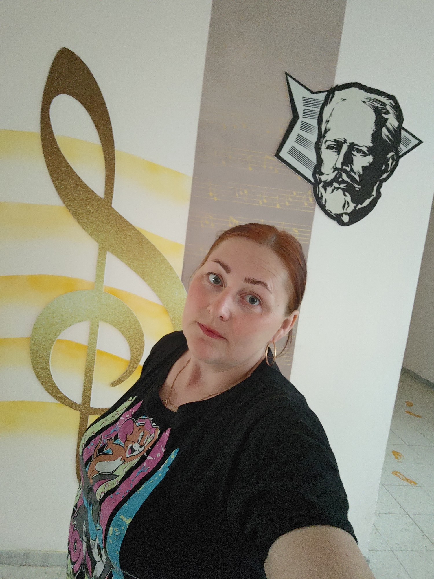 A woman is posing in front of a wall. She has red hair, fair skin, and is wearing gold hoop earrings and a black t-shirt with cartoon characters. Behind her, the wall is decorated with musical motifs: a large, shimmering golden treble clef on a background of white and yellow horizontal stripes, and a black and white portrait of a man with a beard, likely a composer, set against a background of musical notes.
The location is implied to be Markovskii, Russia, based on the surrounding data.