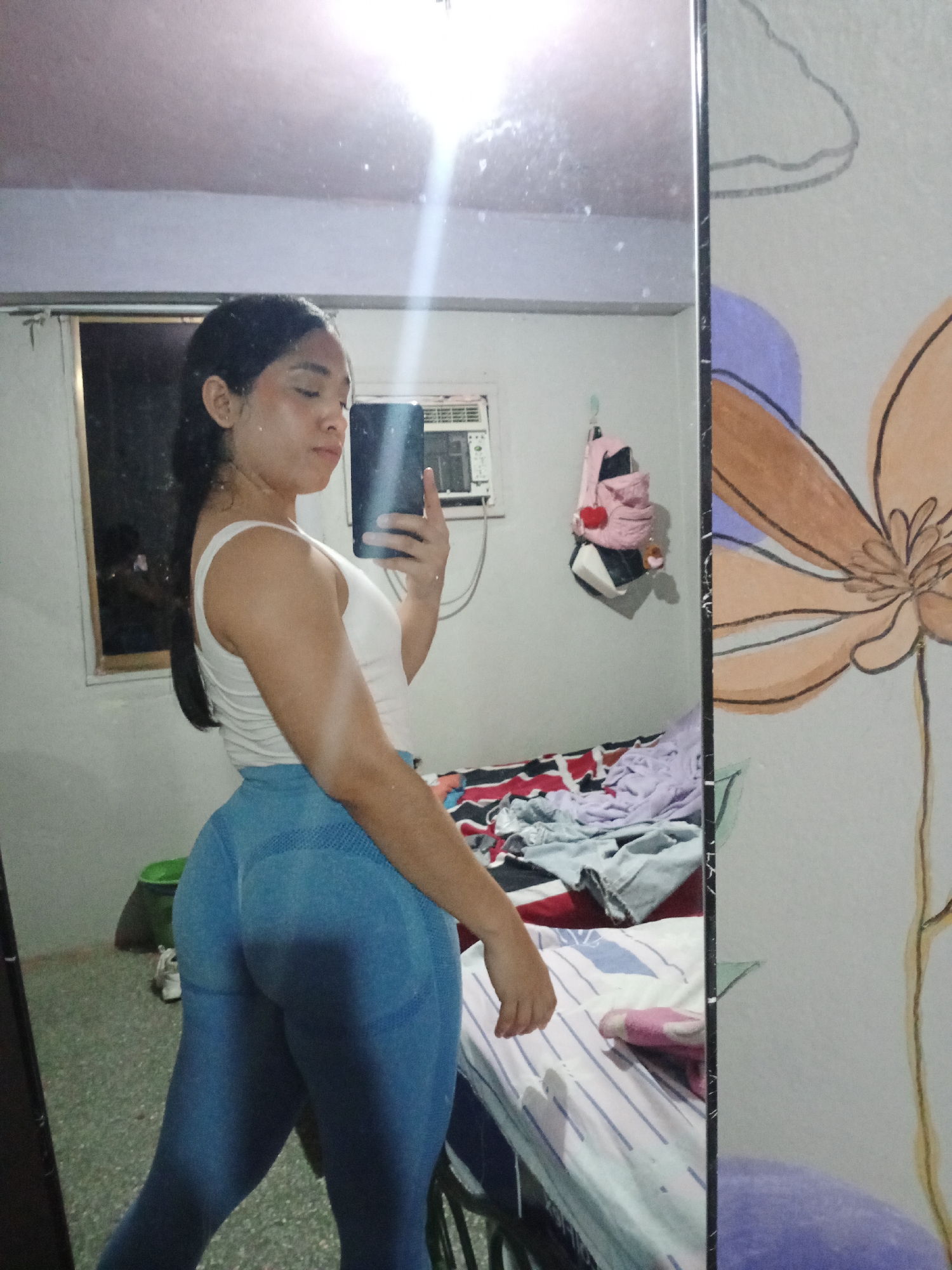 A young woman takes a mirror selfie in an indoor setting. She has dark hair pulled back and is wearing a white tank top and blue leggings. Her phone is visible in her hand. The room appears to be a bedroom, with a bed covered in rumpled bedding and clothing. Other details include a window with bars, an air conditioner, and a backpack hanging on the wall. The room has painted decorations on the mirror. The photo was likely taken in a personal residence. There is no indication of being in the city of Valle de La Pascua, Venezuela, other than a general area where the photo might have been taken.