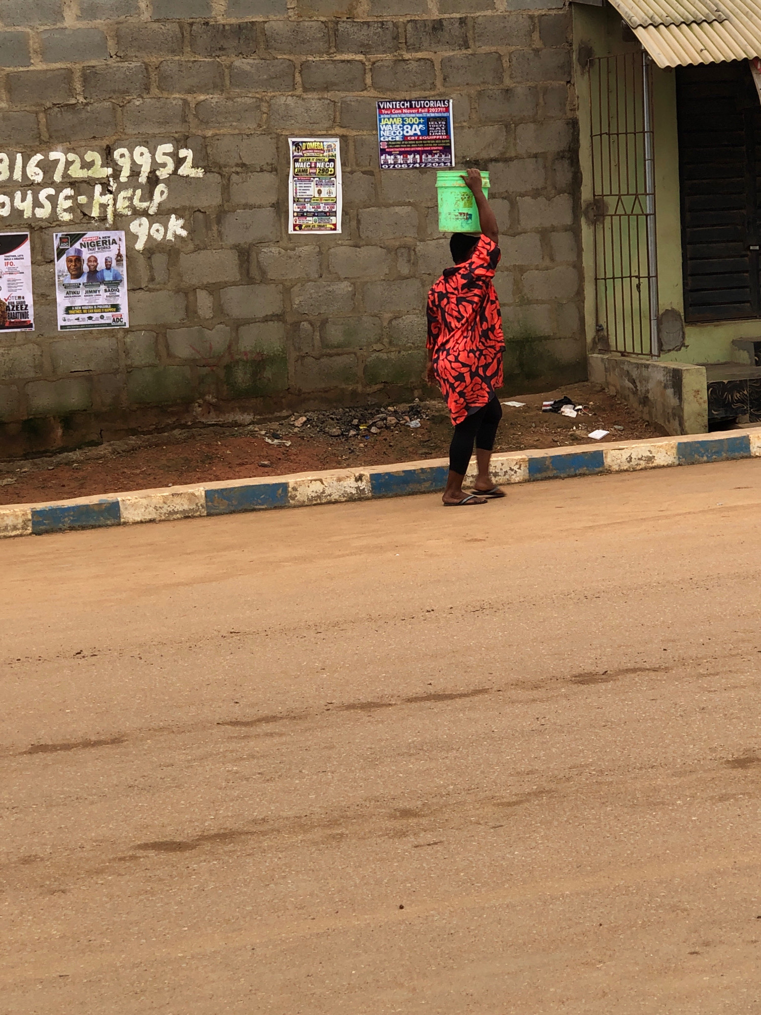 In Lagos, Nigeria, a person wearing a bright red and black patterned shirt and black leggings carries a green bucket of water on their head. They are walking on a dirt road next to a wall covered with various posters and graffiti. The posters advertise educational services like "Vintech Tutorials" and "D'Omega," with some bearing phone numbers and prices. The wall itself is constructed from cinder blocks. A blue and white painted curb separates the road from the wall. In the background, a small, weathered building with a corrugated metal roof and barred windows is visible. Debris and trash are scattered on the ground near the wall. The overall scene depicts a common sight in a developing urban environment, where daily tasks like fetching water are carried out alongside commercial advertising.