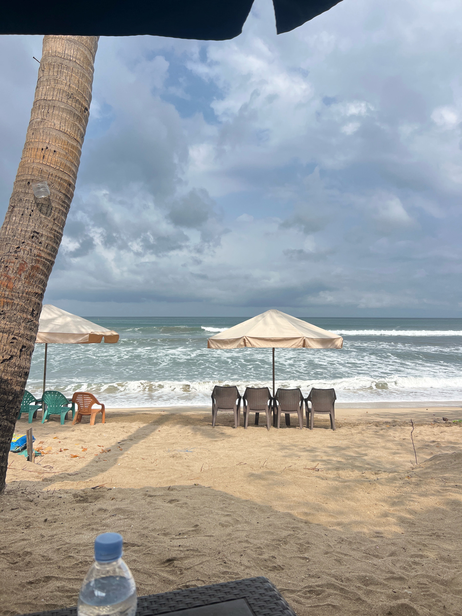 A beach scene in Kuta, Indonesia, under a cloudy sky. In the foreground, a portion of a dark, woven surface is visible, with a partially filled plastic water bottle with a blue cap on it. Further back on the sandy beach are several beach chairs, some green and some orange, positioned near a light-colored beach umbrella. Behind them, another umbrella shades four dark brown chairs lined up. The ocean is visible in the background with moderate waves crashing on the shore. A tall palm tree trunk stands prominently on the left side of the frame. The overall atmosphere is serene, with no people present.