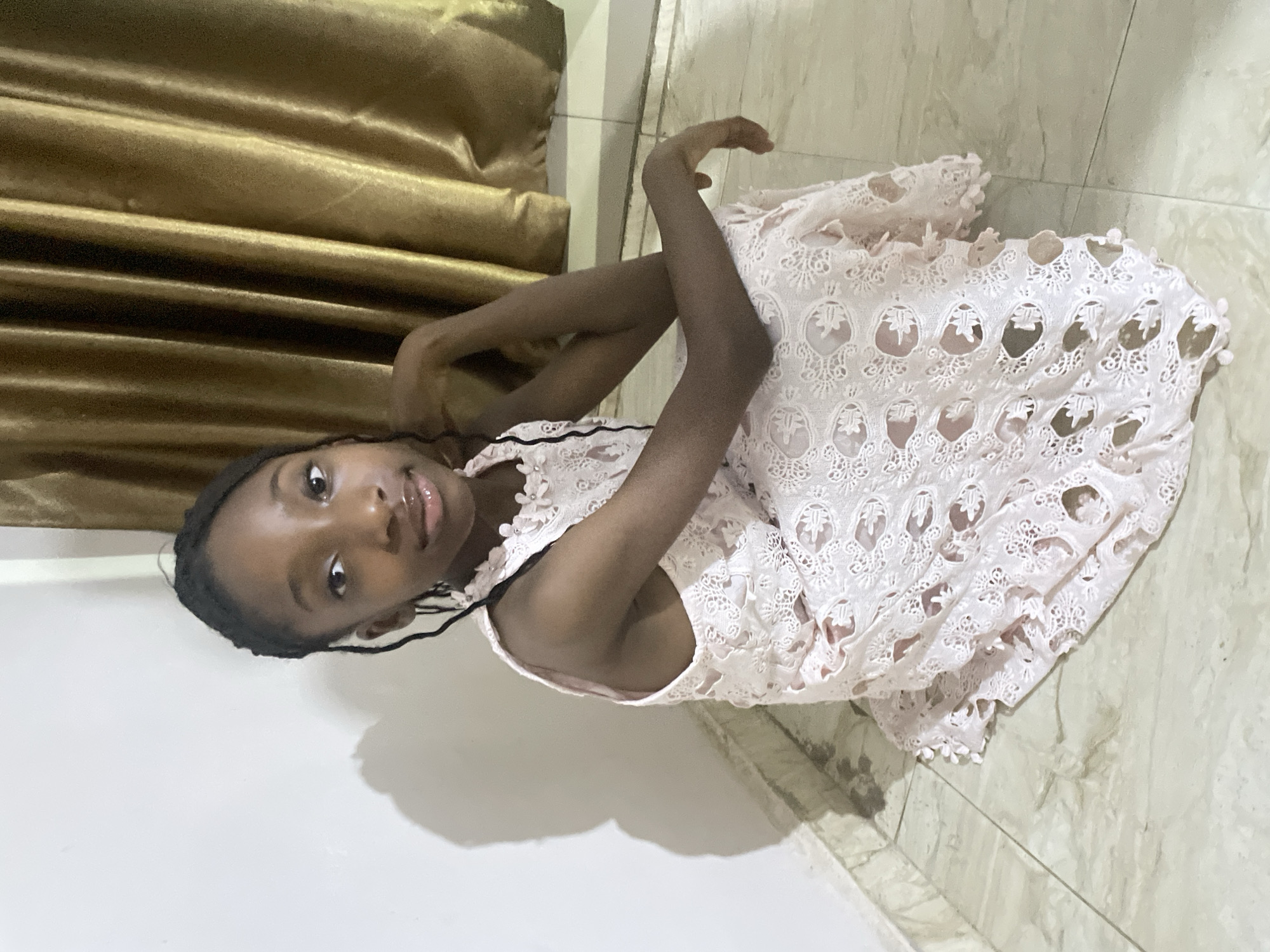 A young girl with dark skin and braided hair lies on a light-colored tile floor. She is wearing a light pink, sleeveless dress with a lace overlay. Her arms are bent, and her hands are near her head. Behind her is a gold-colored curtain or fabric. The scene appears to be indoors, possibly a room in a house. There is no specific information to indicate that this is in Jos, Nigeria.