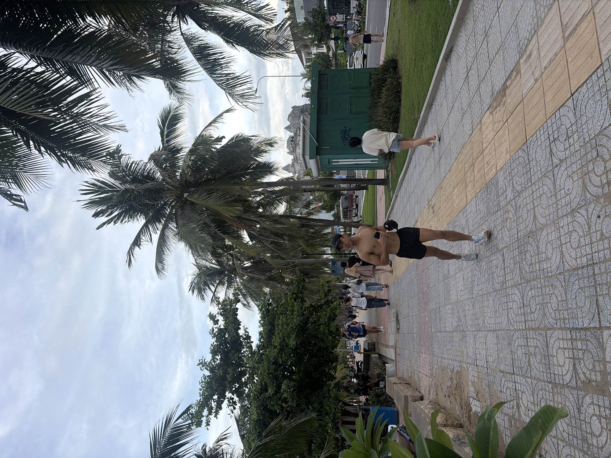 The image captures a scene on a sidewalk in Đà Nẵng, Vietnam, likely near a coastal area given the presence of palm trees. The foreground features a man running in black shorts and white running shoes, holding a dark object, possibly a phone, in his right hand. Behind him, a group of people are walking along the sidewalk. The sidewalk is paved with patterned tiles and a section of tactile paving. To the left of the sidewalk is a grassy area with some low walls and plants. Further back, there is a row of palm trees, and beyond them, a green building with some graffiti, a road with vehicles, and some buildings in the distance, including one with a distinct castle-like structure. The sky is overcast.