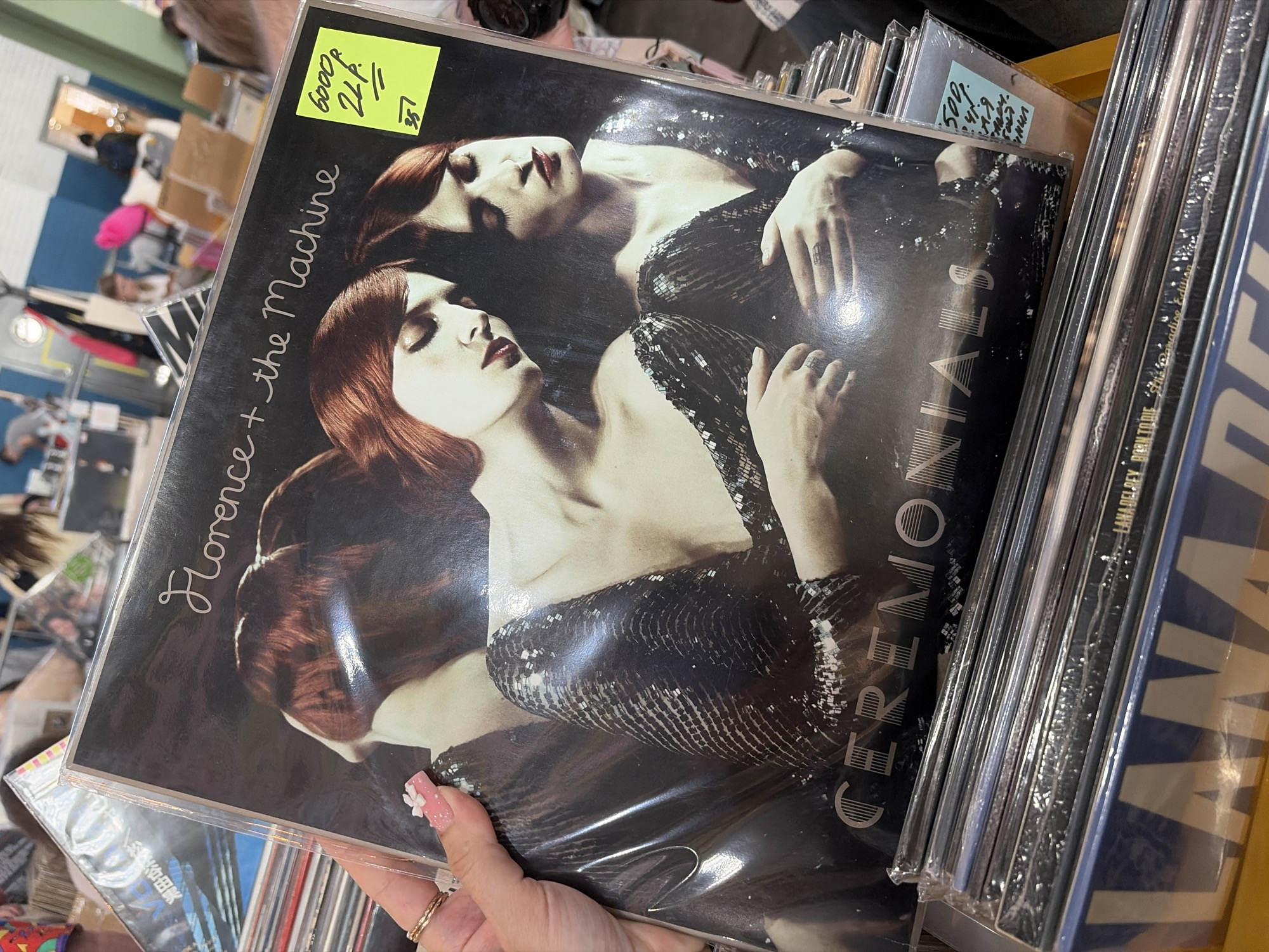 An indoor scene in Saint Petersburg, Russia, depicts a person's left hand holding a vinyl record album. The hand, with light skin tone, wears two gold-colored rings on the ring finger. The thumbnail is manicured with pink polish, white polka dots, and a small white flower appliqué. The record, titled "Florence + the Machine - Ceremonials," is encased in a clear plastic sleeve. Its artwork features two women with reddish-brown hair and dark lipstick, wearing dark, sequined attire, in a close embrace with downcast eyes. A yellow price sticker on the top right corner of the album sleeve displays "6000p," "2LP," and "35" in black handwritten ink. Below the main record, a stack of other vinyl albums is visible in a wooden crate, including one labeled "LANA DEL REY BORN TO DIE The Paradise Edition" and another with a light blue sticker showing "500." The blurred background reveals a spacious interior with sections of blue and white walls, typical of an event hall or large store. Several indistinct figures of people are present, standing and seated. A prominent bright pink object is visible on a surface in the background.