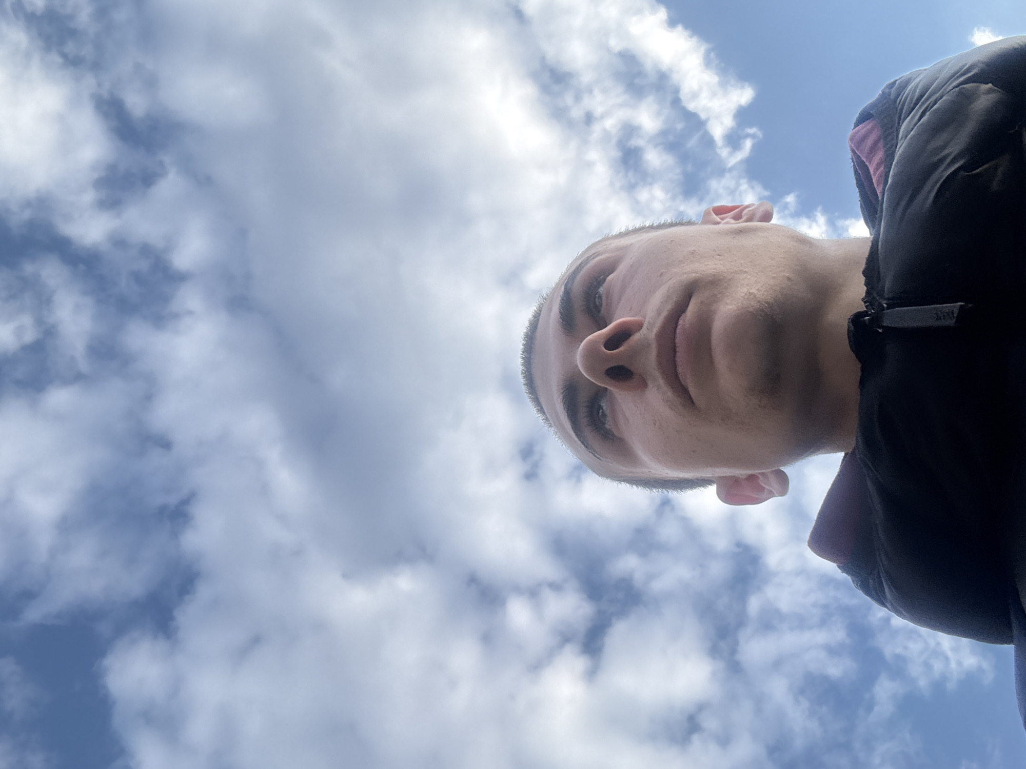 The image presents a low-angle view of a male individual's head and upper torso. The subject, with short dark hair, a prominent nose, and full lips, is positioned in the lower portion of the frame, looking directly upwards. He wears a dark, possibly black, zippered puffy jacket, with a small, indistinct brand marking visible on the zipper pull located on the left side of his chest. A segment of a purple or dark pink garment is visible beneath the jacket. The background is entirely composed of a blue sky with scattered white and light gray cumulus clouds. No specific environmental details or landmarks are discernible to identify a precise location within the city of Plovdiv, Bulgaria.