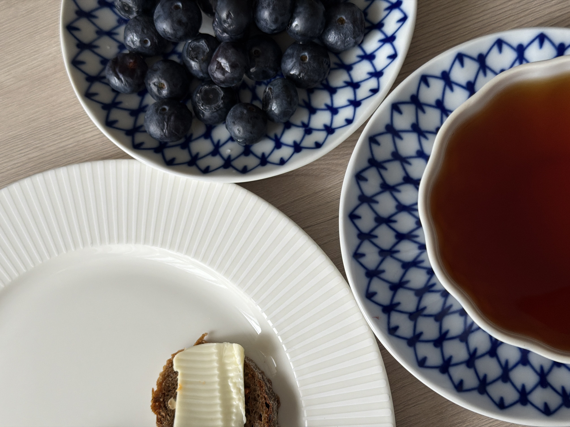A top-down view shows three plates arranged on a wooden surface.
A small, round plate with a blue-and-white geometric pattern holds a pile of blueberries. To the right, a similar patterned plate cradles a teacup filled with a dark liquid, likely tea. To the lower left, a white plate with a vertical ribbed pattern features a slice of bread topped with butter.
The scene suggests a simple breakfast or snack.
There is no information available to link the location to Saint Petersburg, Russia.