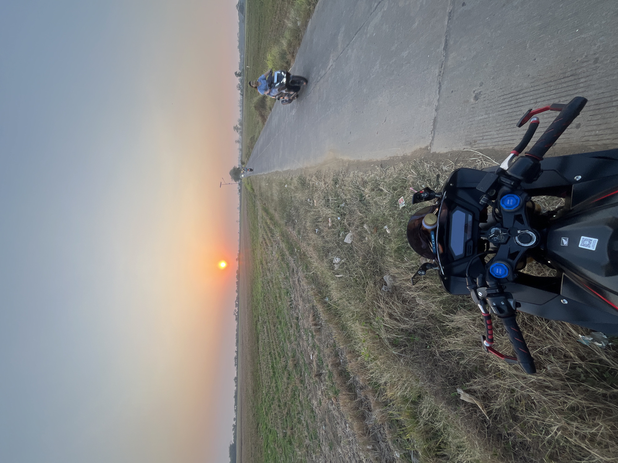 The image captures a serene sunset scene on a rural road in Troso, Indonesia. The foreground is dominated by the handlebars and front portion of a black motorcycle, suggesting the viewpoint of someone on the bike. To the right of the motorcycle, a paved road stretches into the distance, bordered by dry, grassy fields. On the road, two individuals are riding motorcycles; one is closer to the foreground and the other further away. The sky transitions from a clear blue at the top to hues of orange and pink near the horizon, where the sun is setting. The fields appear to be agricultural, possibly harvested, with rows of stubble visible. The overall atmosphere is tranquil, with the natural beauty of the sunset contrasting with the man-made elements of the road and vehicles.