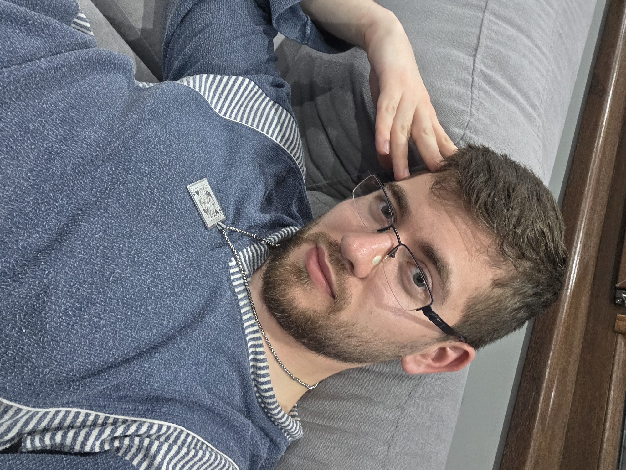 A man with brown hair, a beard, and glasses is lying on a grey surface, possibly a couch or beanbag chair. He is wearing a blue shirt with white stripes along the edges and a playing card pendant necklace. His right hand is near his head. A portion of a wooden frame or door is visible on the right edge of the image. The location in the city of Yagodovo, Bulgaria is undetermined.