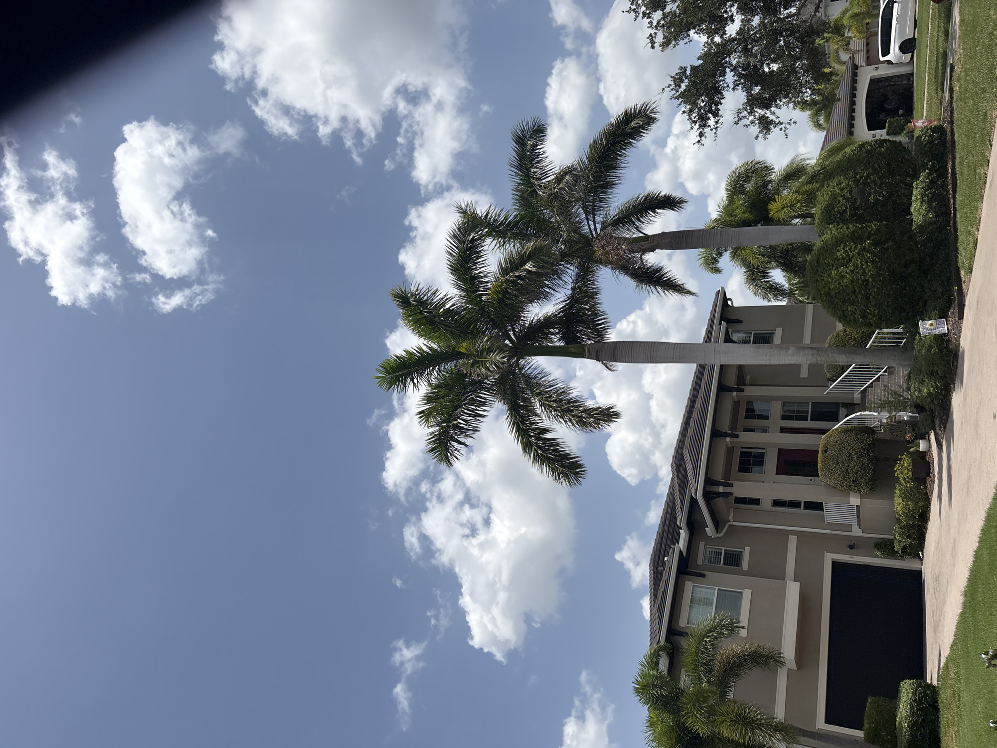 This is a vertical, eye-level, outdoor shot of a modern two-story house in Bradenton, Florida, USA, on a sunny day with scattered white clouds in a blue sky. The house is beige with white trim and features a dark garage door on the left. Two tall palm trees are prominent in the foreground, with their fronds extending towards the sky. Landscaping includes manicured green bushes and grass. To the right, a white SUV is parked in the driveway of a similar-looking house, which has an open garage door revealing some items inside. A partial dark shadow is visible in the upper right corner, suggesting an obstruction to the camera.