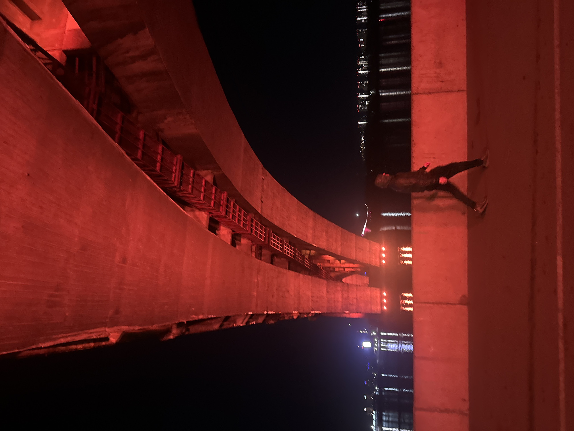 A nighttime, rotated, low-angle image presents a bridge in Krasnoyarsk, Russia. The bridge has concrete walls that curve upward and to the left of the image, with horizontal red metal structures affixed to the concrete walls. Red lighting illuminates these features. A person is seen lying flat, face up, against a section of the bridge’s concrete structure to the right. Distant city lights are visible in the dark background.