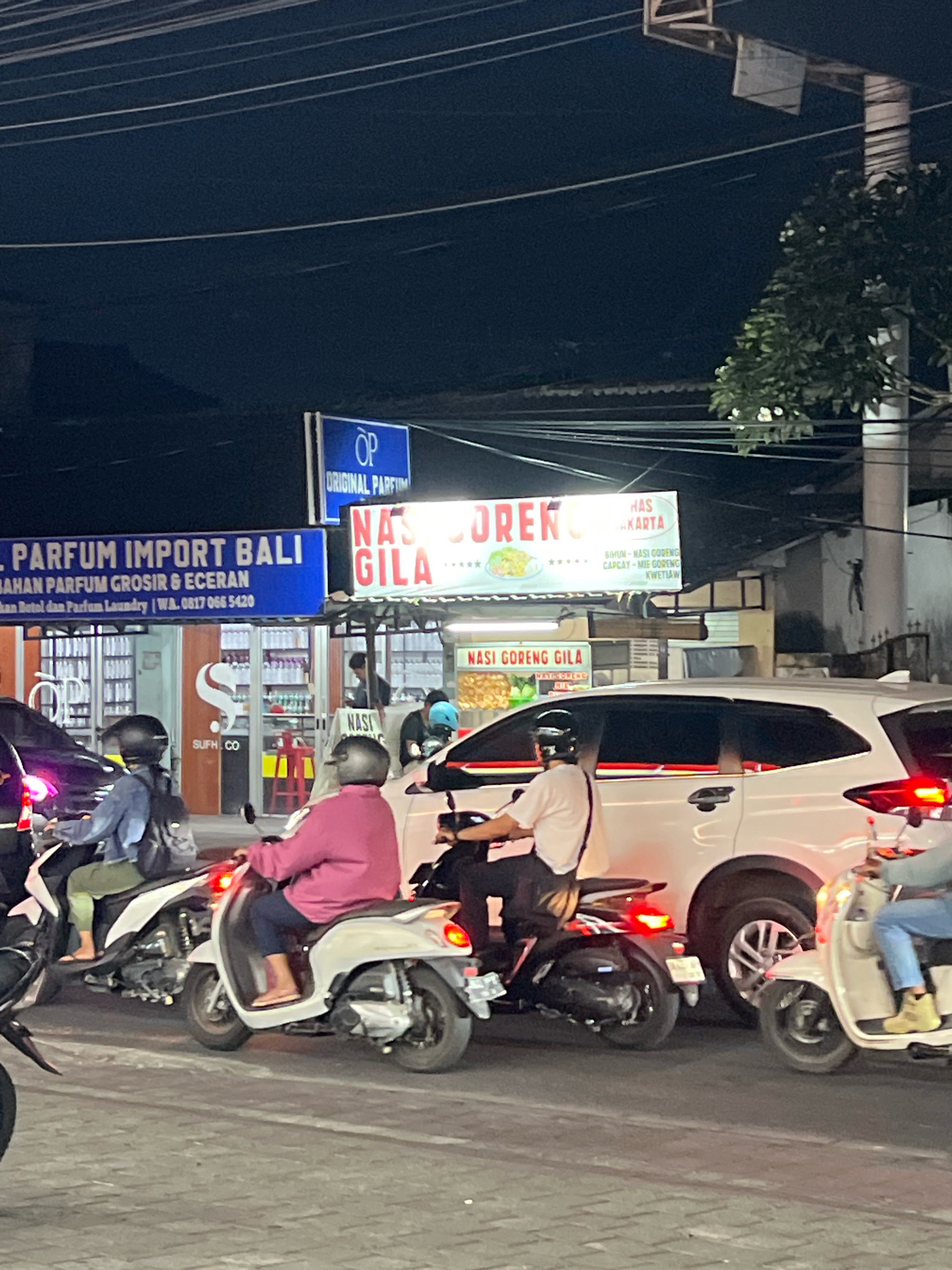 The image depicts a street scene at night in Denpasar, Indonesia, likely at an intersection or busy road. Several motorcycles and one white car are present, indicating traffic. The focus is on the motorcycles, with at least three individuals visible riding them. One person in the foreground wears a pink long-sleeved shirt and blue pants, riding a white scooter. Another rider on a black scooter wears a blue denim jacket and green pants, with a black backpack. A third rider in a light-colored shirt is on a black scooter behind the white car. A white car is stopped or moving slowly in the traffic lane. Overhead, there are power lines and various signs. One prominent sign is illuminated and reads "NASI GORENG GILA" (Crazy Fried Rice), suggesting a food stall or restaurant. Another sign, in blue, displays "OP ORIGINAL PARFUM" and below it "PARFUM IMPORT BALI," indicating a perfume shop. The ground appears to be paved with cobblestones or patterned concrete. The lighting is predominantly artificial, casting shadows and highlighting the illuminated signs. The overall atmosphere suggests a bustling urban environment at night.