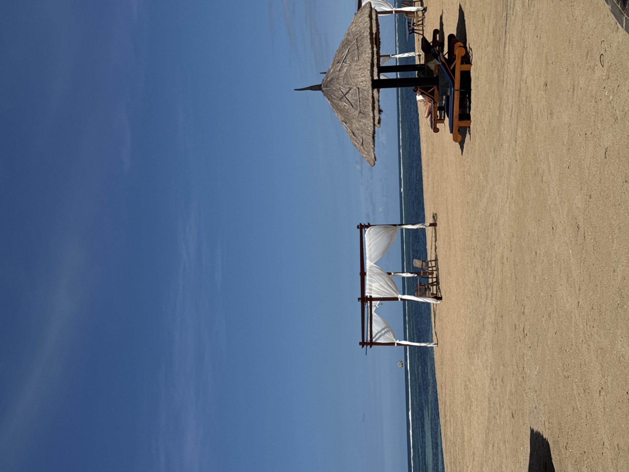 The image captures a beach scene in Kuta Selatan, Indonesia, featuring a blue sky, ocean, and sandy beach. A thatched-roof structure with a black frame is visible on the right side of the image, under which people are sitting on lounge chairs. Further down the beach towards the left, there is a white canopy with a dark frame. The shoreline is seen separating the ocean and beach. The beach occupies a large portion of the right side of the image, with the sky dominating the left side.