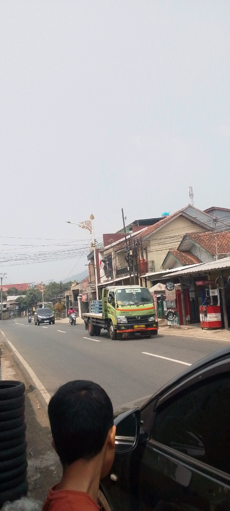 The image captures a street scene in Sukasari, Indonesia, with a focus on vehicular traffic and roadside establishments. In the foreground, a person's head and shoulders are visible, looking towards the right side of the frame, suggesting they are inside or near a vehicle. To the left, a stack of black tires is piled on the side of the road.
The main subject of the image is a green flatbed truck driving on the road. The truck carries several large bottles of water in its bed. It has a license plate that reads "F 9581 CA". Other vehicles are also present on the road, including a black car further ahead and a person on a motorcycle.
The background shows a street lined with buildings. Some of these appear to be shops or businesses, with awnings and signage visible, including what looks like a Pertamina gas station. Power lines crisscross above the street, and in the distance, hills or mountains are faintly visible against a hazy sky. The architecture of the buildings suggests a tropical or Southeast Asian setting. The overall atmosphere is that of a typical day in a developing urban or semi-urban area.