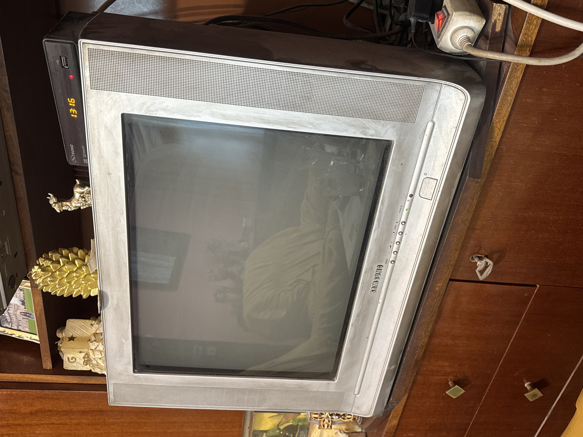 The image shows a silver Samsung CRT television sitting on a dark wooden cabinet. A black set-top box is on top of the TV, displaying "13:15" on its digital display. Various power cables and a white power strip are visible behind the television. On the shelf to the left of the TV, there are gold pineapple-shaped objects and a framed picture. A reflection of the room is visible on the TV screen. The location cannot be determined from this image alone. The location in the city of Plovdiv, Bulgaria is unknown.
