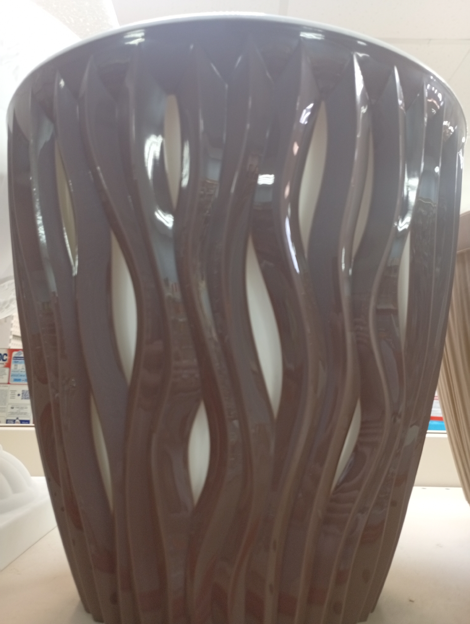 A close-up shot shows a decorative, brown, vase-like object with a wavy, openwork design. The vase is glossy and reflects light, highlighting its curved surfaces and the empty spaces between the vertical, flowing elements. Inside the vase, a lighter color, possibly white, is visible. The background is out of focus, with hints of a bright, possibly ceiling, light source above. To the left of the vase, there are parts of what appear to be boxes or labels with text and graphics, some of which are partially obscured. To the right, a beige, fabric-like material hangs vertically. The object is situated on a flat, light-colored surface. The scene suggests a retail or display setting. This image was captured in Borovichi, Russia.