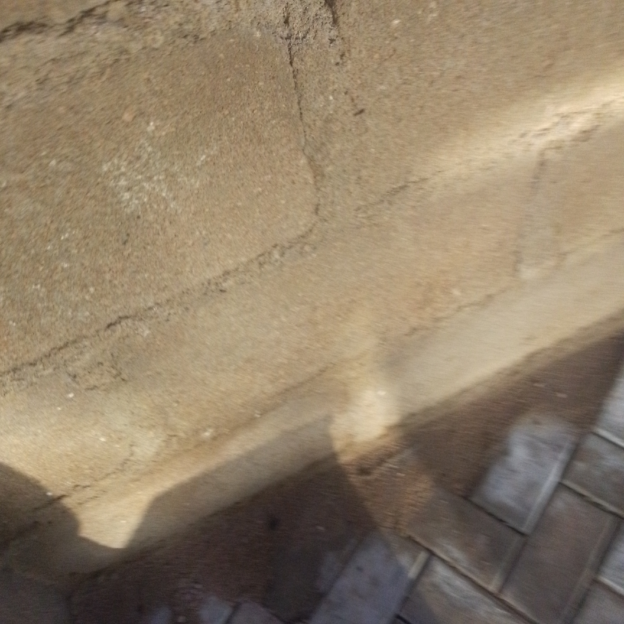The image depicts an outdoor scene with a textured, sandy-colored wall in the upper portion, possibly made of concrete blocks. A dark shadow stretches across the lower part of the image, cast by an unseen object or person. The ground in the lower right appears to be paved with interlocking bricks, laid in a herringbone pattern. The lighting suggests it is daytime, with the shadow indicating a strong light source, likely the sun. The scene appears to be in Jalingo, Nigeria, as indicated by the location context. There is no visible text in the image, and the image is quite blurry, making it difficult to discern specific details or emotions.