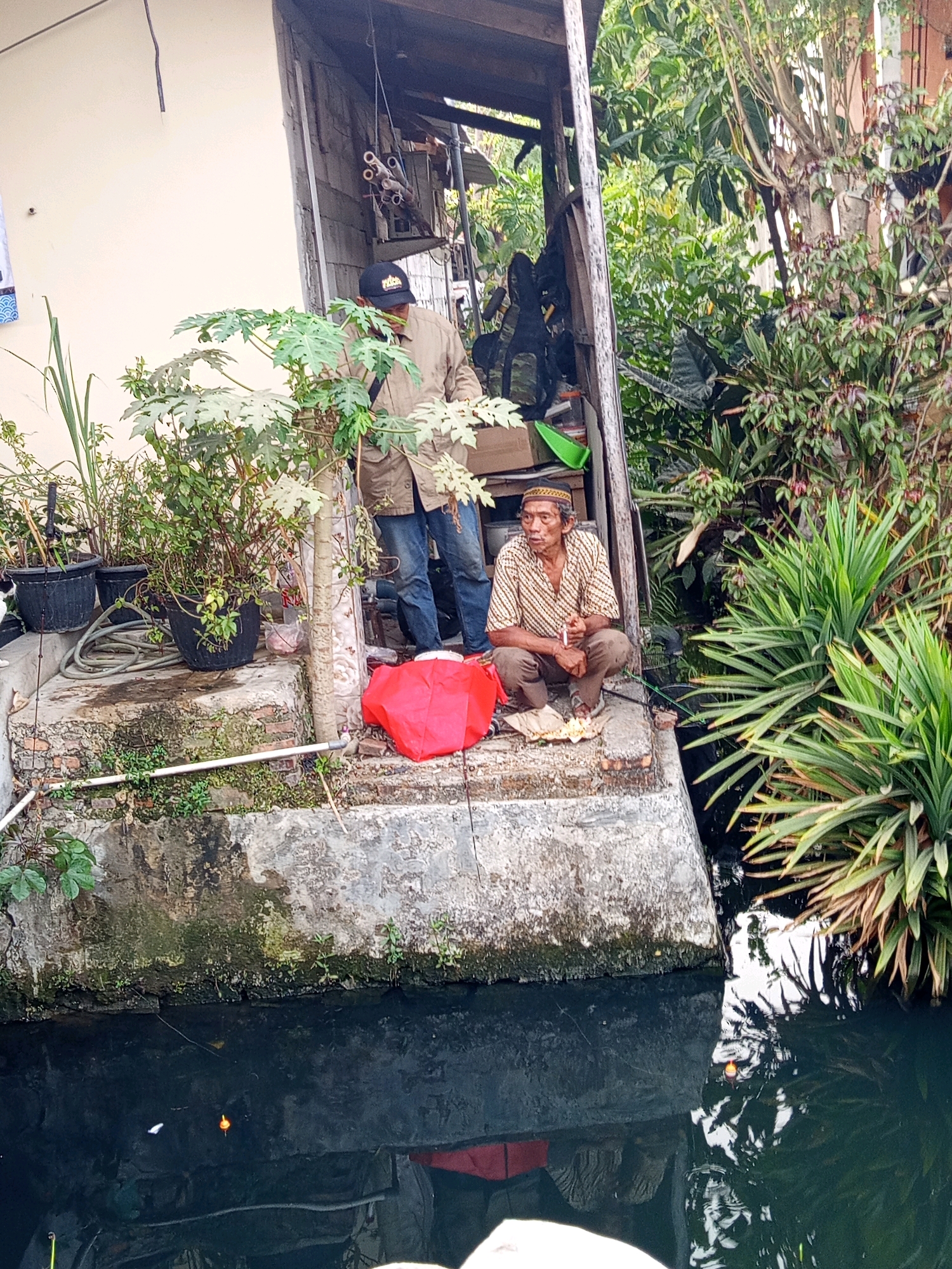 In Karawang, Indonesia, an outdoor scene unfolds where two men are present near a body of dark, still water. The man on the right, who is older and wearing a striped shirt and a tan cap, is crouched down and holding a cigarette. He is positioned on a concrete structure adjacent to the water, next to a red bag. To his left, another man stands wearing a light-colored jacket and blue jeans, also near the concrete structure, with a backpack slung over his shoulder. The environment is overgrown with lush green foliage, including large-leafed plants and trees, surrounding the area. The concrete structure appears weathered and has plants growing on its edges. In the water, fishing floats are visible, indicating potential fishing activity. The overall impression is of a humble, somewhat rustic environment within a city.