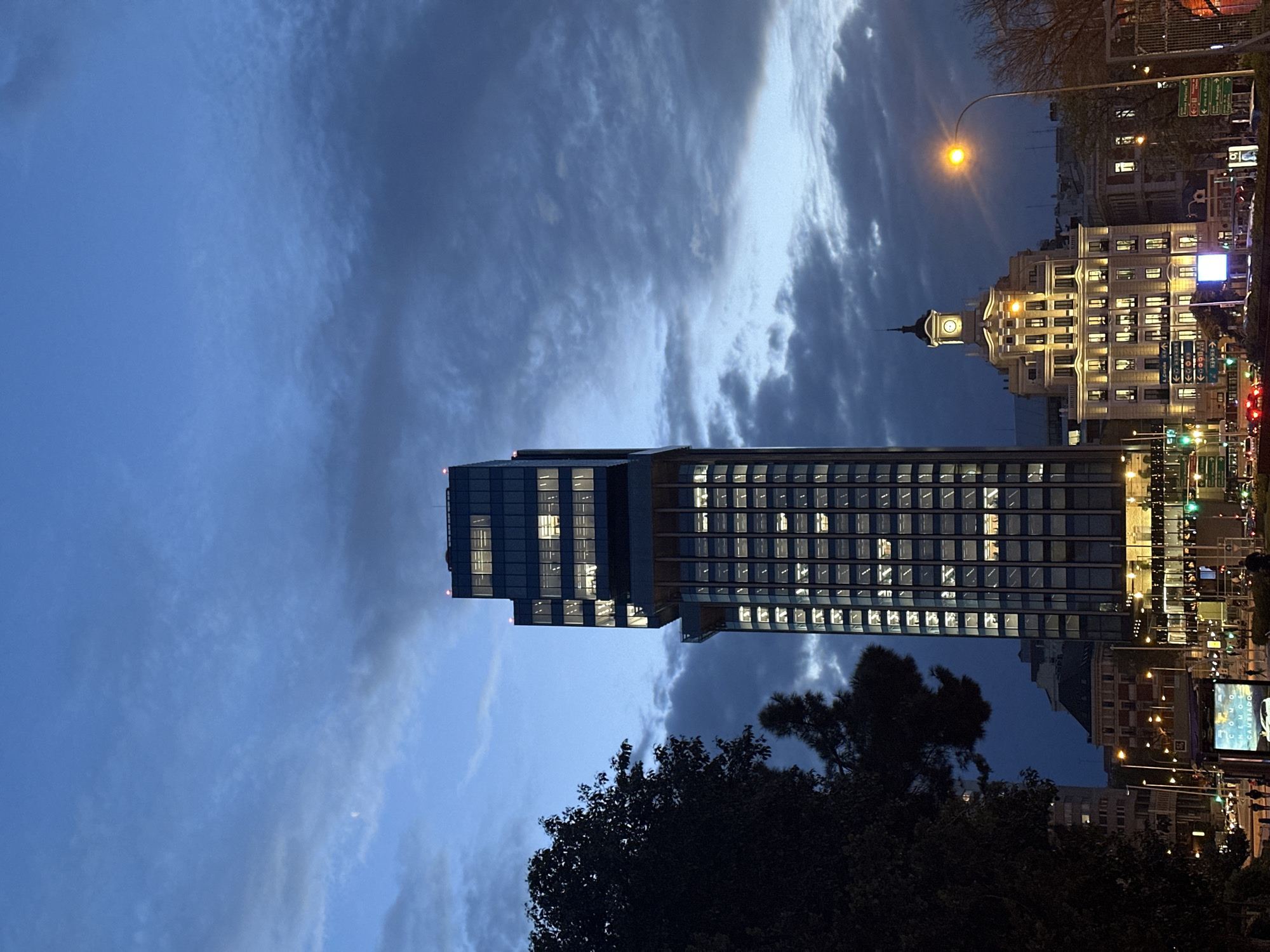 The image shows a nighttime urban landscape in Madrid, Spain. Prominently featured is the Torre Picasso skyscraper, a modern, rectangular structure with numerous illuminated windows. To the right of the Torre Picasso is a more ornate, classical building with a clock tower, also brightly lit. A street lamp casts a warm glow over the scene. Traffic lights and vehicle lights suggest active traffic flow. The lower part of the image includes silhouetted tree foliage. The sky is filled with dark, textured clouds.
