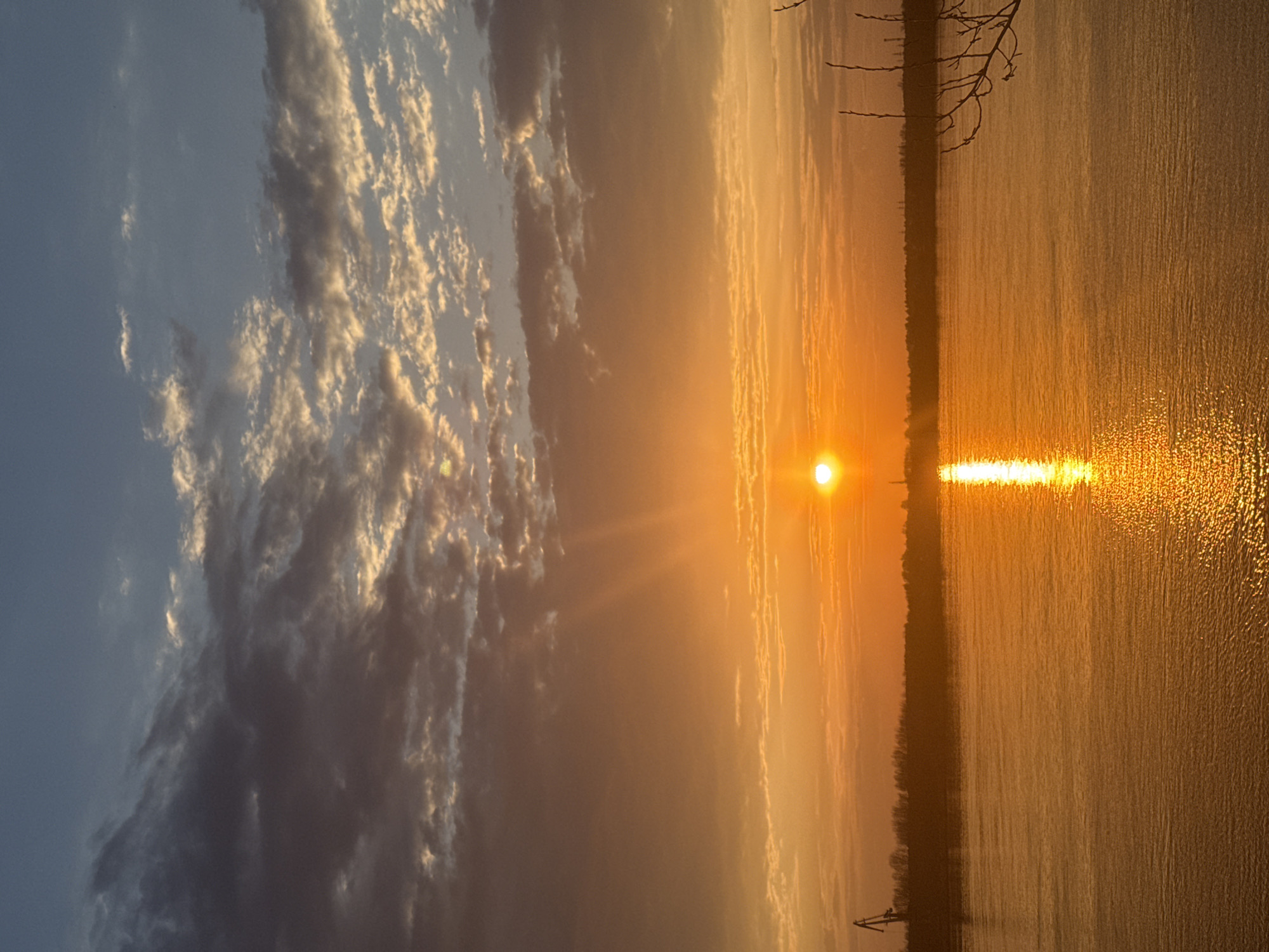 The image depicts a sunset over the Kama River in Chaikovsky, Russia. The sun is positioned near the horizon, casting a bright reflection on the water's surface. The sky on the left is a mix of blue and cloudy formations, while the sky closer to the sun is tinged with orange and yellow hues. The opposite shore is visible in the distance, marked by a line of trees. A few bare tree branches extend into the upper right corner of the frame. The river's surface shows subtle ripples and variations in light, due to reflection of the sun.