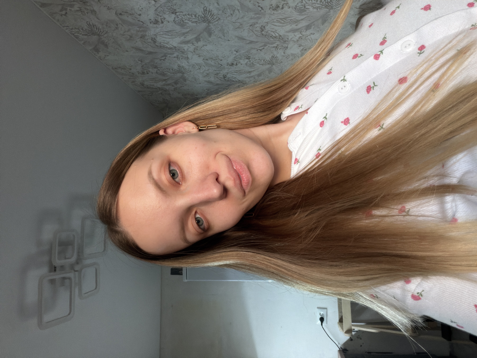 A woman with long blonde hair is posing for a selfie. She is wearing a white shirt with small red floral patterns. Her hair is partially covering her face and shirt. She is wearing a gold earring. The background includes a patterned wall, a plain wall, and what appears to be a power outlet near the bottom. The location is in Reshetnikovo, Russia.
