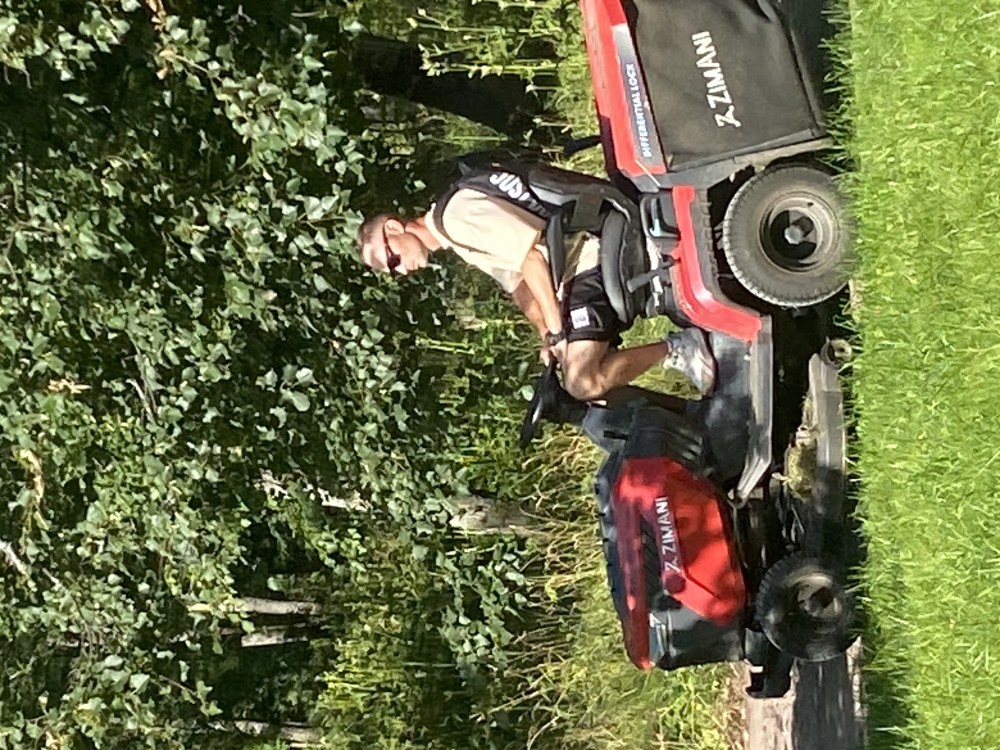 A man is riding a red and black riding lawn mower in a grassy area. He is wearing sunglasses, a light-colored t-shirt, black shorts, and gray athletic shoes. He has a black backpack on with white lettering. The lawn mower has a grass catcher bag attached to the side with "ZIMANI" printed on it. The background consists of lush green trees and vegetation, suggesting an outdoor, possibly rural or park-like setting. The image does not provide information about the specific location within the city of Pushkin, Russia.