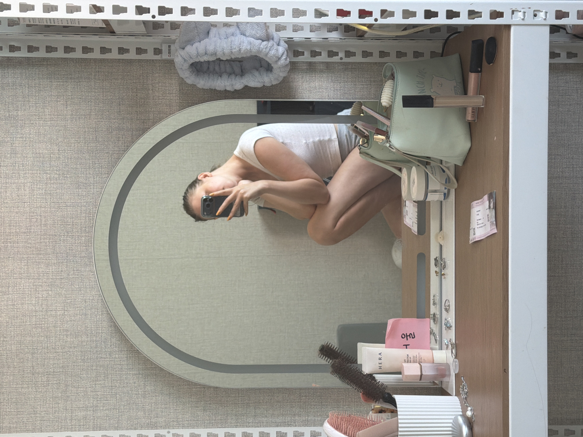 A young woman with blonde hair pulled back is photographed in a mirror selfie. She is crouched down in front of a vanity desk, wearing a white t-shirt and grey shorts. She holds a black smartphone in her right hand, taking the picture. The desk is cluttered with various beauty products, including a light green makeup bag, tubes of lotion, brushes, rings, and what appear to be small jars. A pale grey, fluffy headband hangs on a white shelf unit to the right of the mirror. The background is a textured, light grey wall. The setting is likely a personal vanity area, possibly in a bedroom or dressing room. The image does not contain any identifiable landmarks or context to suggest a specific location within Dongducheon-si, South Korea, beyond it being an indoor scene.