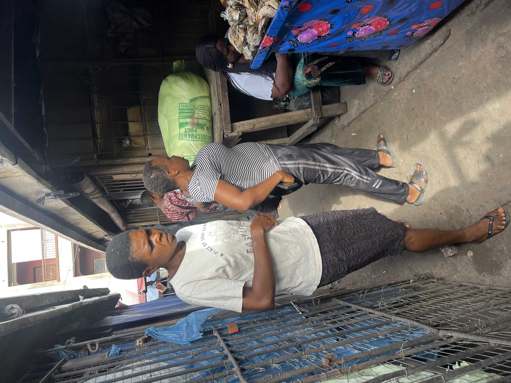 The image depicts an outdoor scene in an urban environment, possibly a market. In the foreground, a person wearing a white t-shirt and dark skirt lies on what appears to be a metal grate. Behind this person, another individual in a striped shirt and track pants is also lying down. In the background, a woman sits on a wooden structure, possibly selling goods. A green sack is beside her, and a blue floral fabric drapes over the structure. The overall scene suggests a resting moment amidst a busy commercial area.