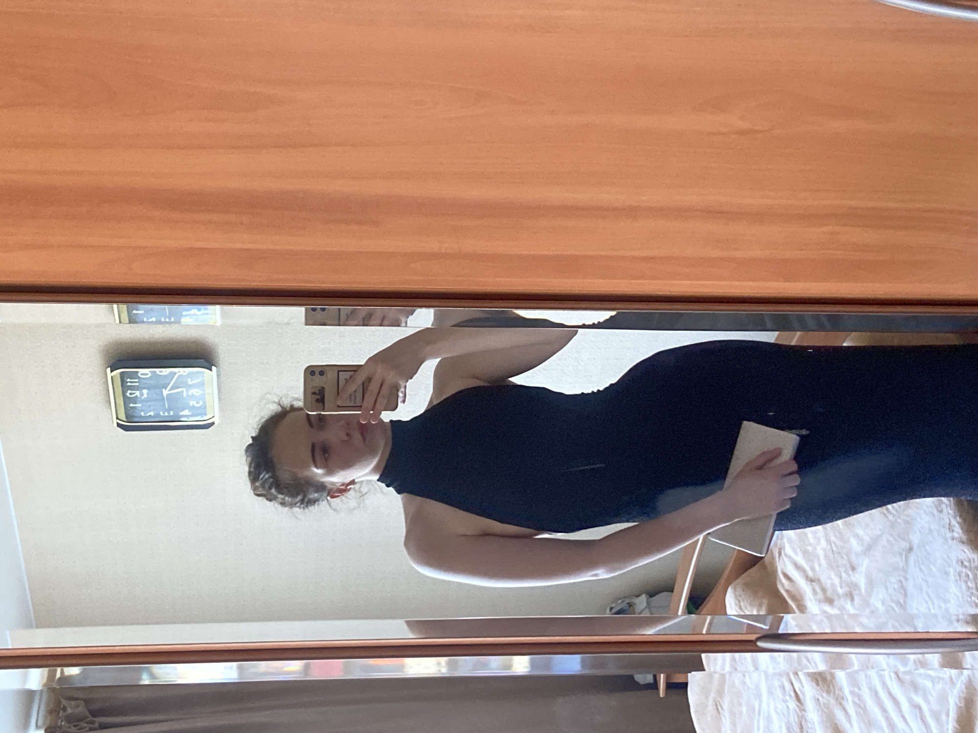 A person is captured in a mirror selfie, wearing a black dress. The setting appears to be indoors, possibly a bedroom, with a wooden-paneled wall or furniture reflecting in the mirror. A clock on the wall shows it is past 1 PM, and the lighting suggests it is daytime. The person is holding a smartphone, presumably to take the photo, and another hand is holding a light-colored rectangular object. The reflection also shows a beige bedspread. The image is oriented sideways. There is no visible text in the image.