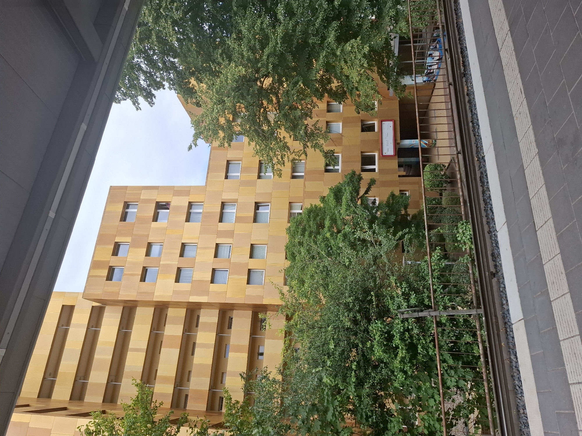 The image captures a view of the residential buildings at the "Flugfeld" residential complex in Hamburg, Germany. The buildings are characterized by their distinctive golden-yellow facade, composed of square panels. The architecture features multiple stories with numerous windows and balconies. Lush green trees are interspersed with the buildings, partially obscuring some of the lower levels. In the foreground, there are train tracks and a platform, indicating the presence of a train station or stop. The overall scene suggests an urban environment with a mix of residential architecture and natural elements.