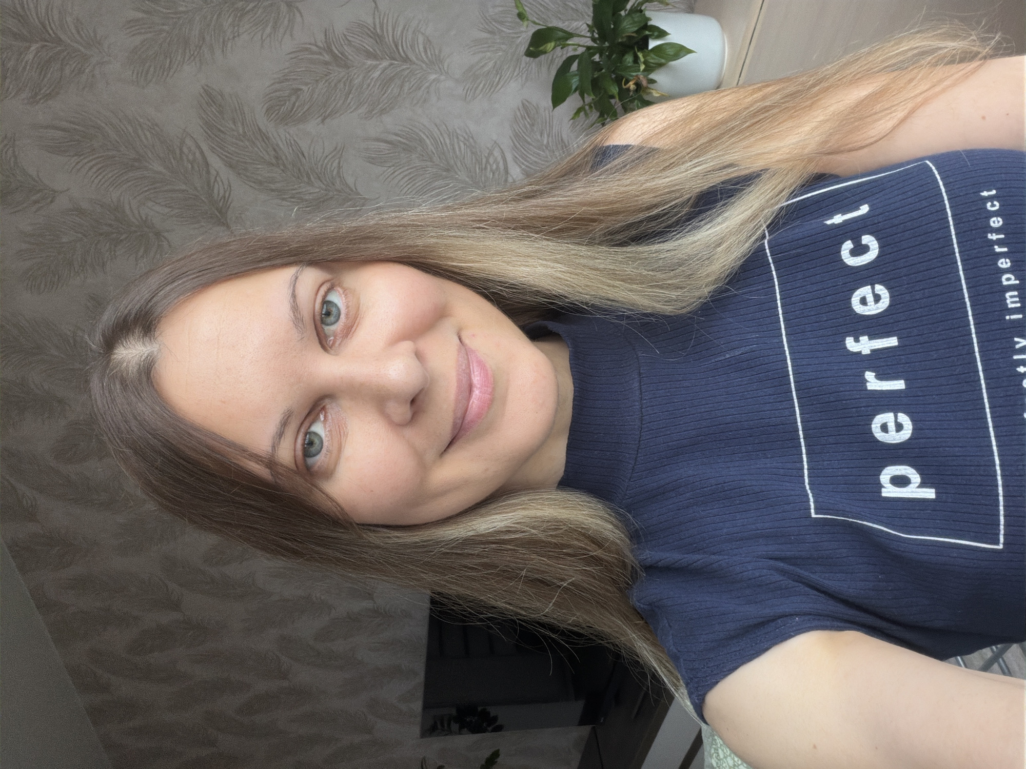 The image shows a medium shot of a woman with long, blonde hair and blue eyes. She is wearing a dark blue, sleeveless, ribbed top with the word "perfect" printed in white text within a white rectangle. She is smiling gently and looking directly at the camera. The background consists of a textured wallpaper with a feather-like pattern and a potted plant on the right side of the frame. To the left, a television screen is visible. The lighting suggests an indoor setting, possibly a home or apartment. There is no indication of the specific location within the city of Izhevsk, Russia, beyond it being an indoor scene.