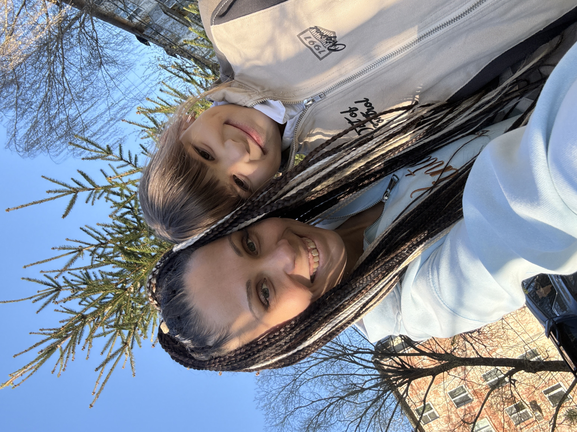 An outdoor selfie captures two individuals, an adult female and a younger person, smiling cheerfully in Rusanovo, Russia. The image is taken from a low-angle perspective against a bright blue sky, with the subjects appearing rotated ninety degrees clockwise.
The adult, in the foreground, has dark braided hair with distinct white or light-colored streaks. She is wearing a light blue hooded top, featuring the partial text "Heart of gold" in a stylized, copper-colored font. Her eyes are greenish-brown, and she displays a broad, happy smile.
Behind her, the younger person also smiles towards the camera, revealing lighter brown, straight hair with bangs. They are dressed in a light beige jacket over a white collared shirt. Visible on the jacket are a square logo containing "1991" and the stylized text "Old School."
The background consists of a clear, bright blue sky, indicating sunny daytime conditions. A lush evergreen tree branch is prominent in the upper left, while bare branches of deciduous trees partially frame a multi-story brick building with windows in the lower right. The bright light casts gentle shadows, highlighting the cheerful expressions of both individuals.