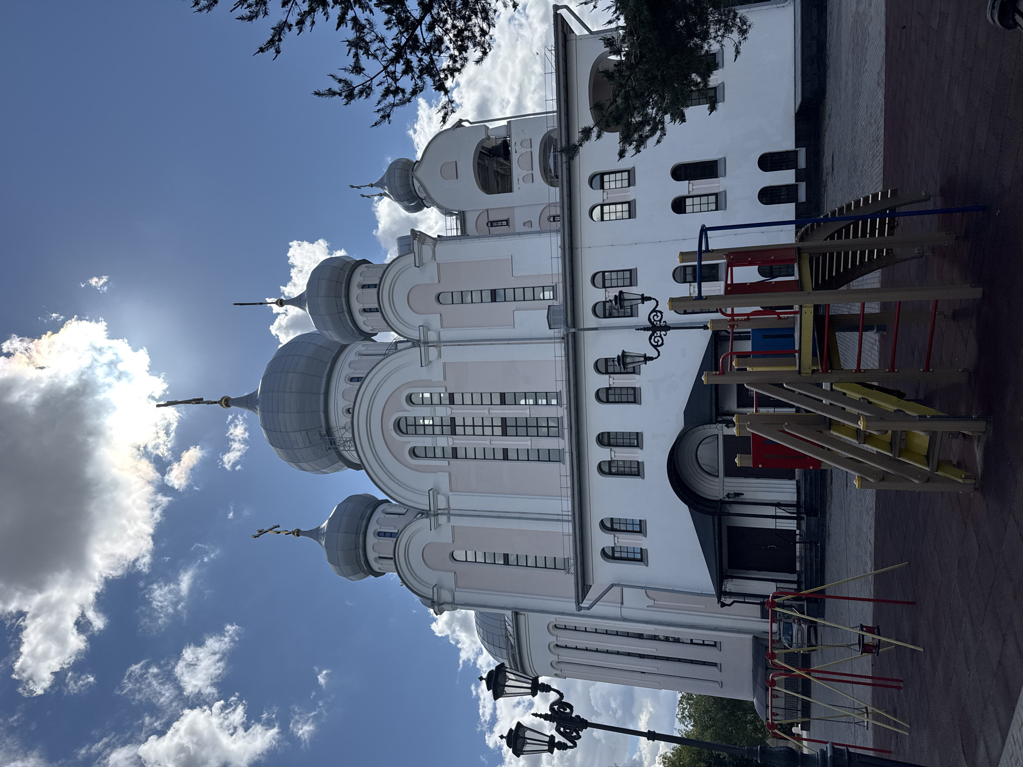 The image depicts a white church with pink accents and multiple gray domes, topped with crosses. In the foreground, there is a playground with swings and a slide. A street lamp stands to the left of the church. The sky is blue with scattered white clouds, and the sun is visible behind one of the clouds. The church is located in Saint Petersburg, Russia.