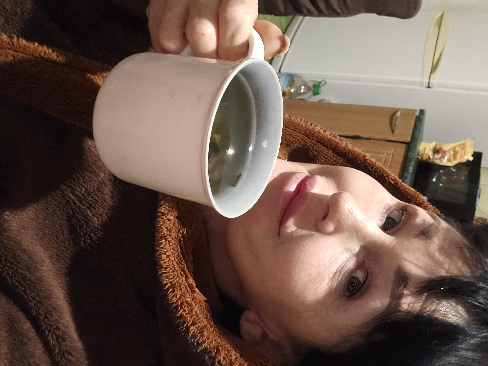 An older woman with dark hair is depicted indoors, looking directly at the camera with a slight smile. She is wearing a brown, textured, possibly fuzzy robe or blanket that covers her shoulders and chest. Her right hand holds a white mug containing a light green liquid with visible floating particles. In the background, to the left, a white refrigerator is visible. To the right, a kitchen counter with a light green surface and light brown wooden cabinets with a silver handle can be seen. On the counter, a yellow squeeze bottle and a dark appliance are present. A clear plastic bottle with a green cap is partially visible near the floor. This scene is situated in Nova Kakhovka, Ukraine.