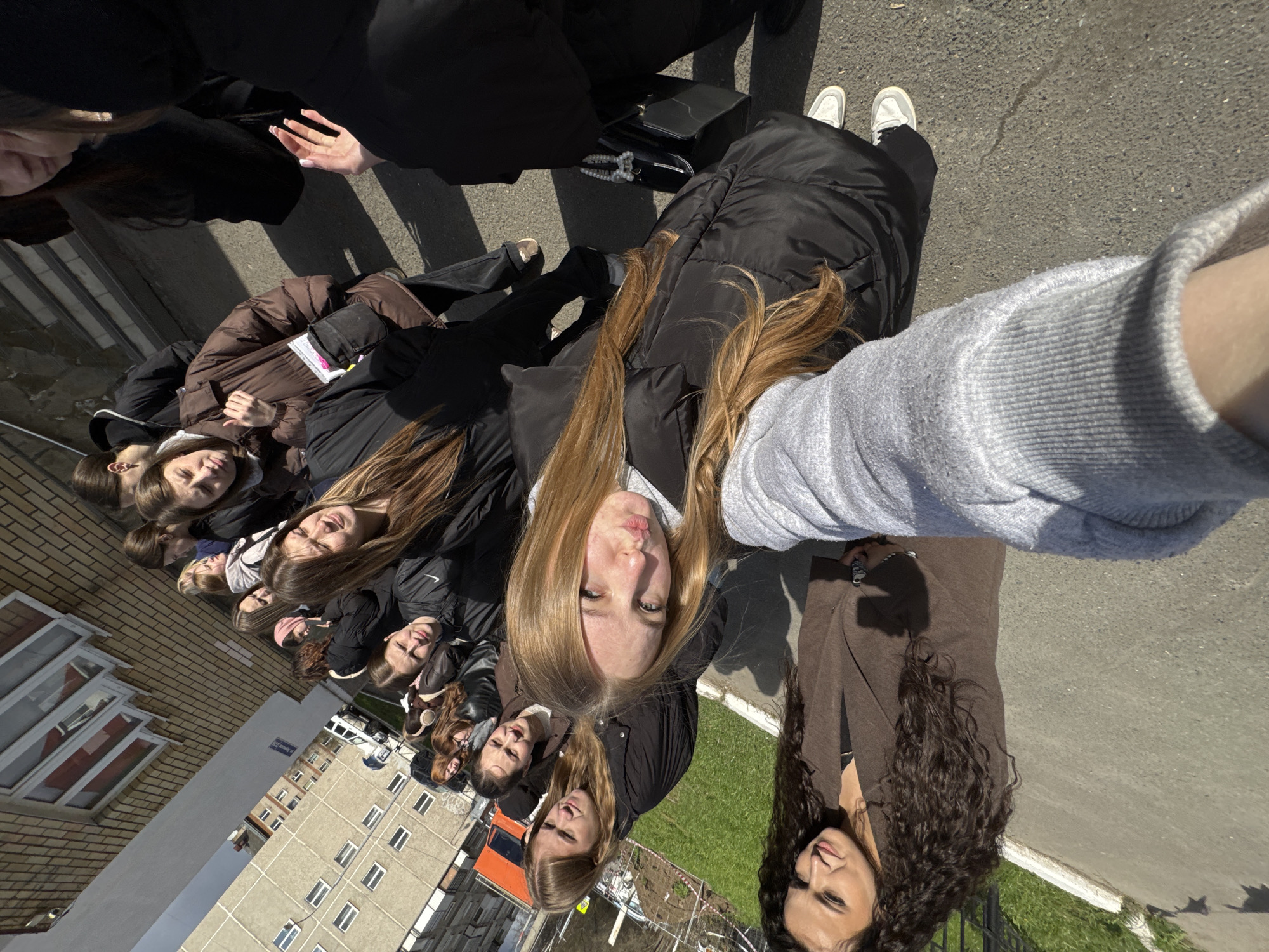 This media file captures a high-angle group selfie of approximately ten young women outdoors on a bright, sunny day in Perm, Russia. The image is taken from a top-down perspective, with the arm and grey long-sleeved top of the person holding the camera visible in the immediate foreground.
Most of the women are looking upwards towards the camera, their faces illuminated by direct sunlight, creating distinct highlights and shadows. Their expressions range from neutral to slight smiles, with one woman with long blonde hair looking directly at the camera with a subtle pout. Another woman with long, dark, curly hair is partially visible on the far right, also looking up. They are dressed in various winter or cool-weather attire, including black puffer jackets, a brown puffer jacket, and other dark coats. Visible accessories include a black handbag with a pearl-like strap resting on the ground near one person, and several pairs of sneakers, including white ones.
The setting is an urban outdoor space. The ground is predominantly asphalt with patches of green grass. In the background, several buildings are visible: a multi-story brick building with rows of windows to the left, and a larger concrete building with numerous windows further back and to the right. A white sign with blue, unreadable text is affixed to the concrete building. Fences are also partially visible. The clear sky and strong shadows indicate bright sunlight, suggesting mid-day to afternoon. The architecture of the buildings is consistent with the specified location of Perm, Russia.