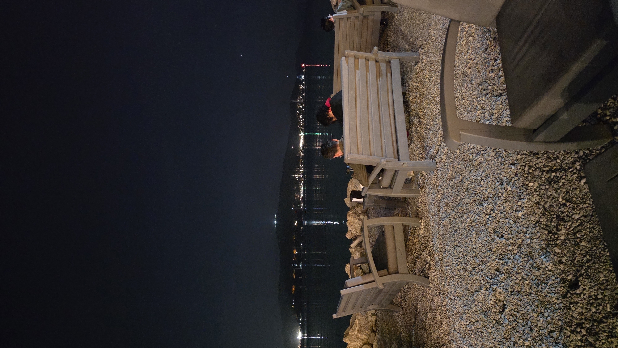 This nighttime photograph, taken in Fethiye, Turkey, captures a serene waterfront scene. The image, when viewed with a level horizon, shows a dark sky at the top, a body of water in the middle, and a gravel-covered area with benches in the foreground towards the bottom right.
In the immediate foreground, the ground is covered with numerous small, light-colored gravel or pebbles, forming a pathway or seating area. Several simple, light-toned wooden benches, featuring slatted seats and backs, are arranged facing the water. Three individuals are visible seated on these benches, all with their backs to the viewer, engrossed in the view. The person closest to the camera wears light-colored clothing and has dark hair. On a slightly more distant bench, another individual with dark, curly hair has a noticeable red element on their clothing, possibly a hood or collar. A third person on the furthest visible bench is largely obscured.
The midground features a calm body of water, likely a bay or lake, which beautifully reflects the numerous artificial lights from a distant settlement. These lights stretch horizontally across the background, outlining a town or city along the opposite shore or base of distant mountains. The sky above is a deep, dark navy or black, with one particularly bright star or planet visible towards the upper left.
The overall scene conveys a peaceful and contemplative atmosphere, with people quietly enjoying the illuminated night vista from the comfortable vantage point of the benches. There is no visible text in the image.