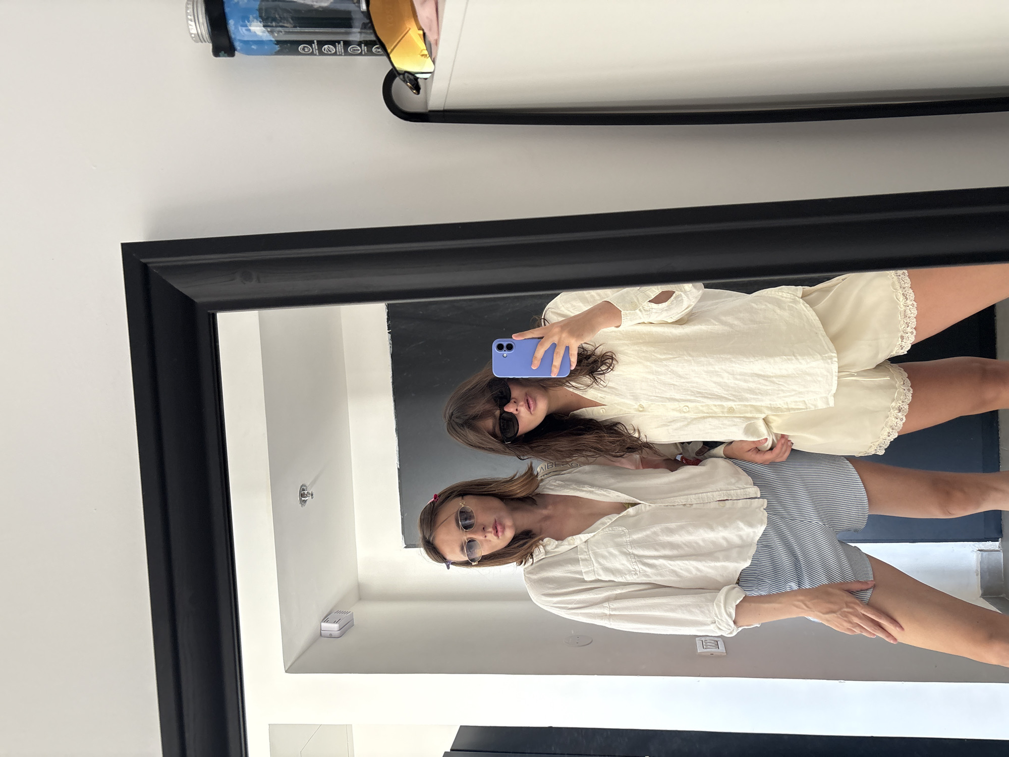 This image is an indoor mirror selfie taken by two women in a modern room, likely an apartment or hotel, in Tel-Aviv, Israel.
The woman on the left, standing slightly behind and to the left of the woman taking the photo, has shoulder-length brown hair with a red and a blue hair clip visible. She wears small, oval-framed silver sunglasses, a loose-fitting white long-sleeve button-down shirt with some buttons undone, and blue and white striped shorts. Her right hand is resting on her left upper thigh.
The woman on the right, taking the photograph, has long brown hair and wears dark, rectangular-framed sunglasses. She is dressed in a loose-fitting cream-colored long-sleeve button-down shirt and matching cream-colored shorts with white lace trim along the hem. She holds a purple-cased smartphone with both hands, partially obscuring the lower half of her face as she photographs their reflection in the large, black-framed mirror that dominates the center of the frame.
The reflected background shows a predominantly white room with light-colored walls and a matching floor. Visible architectural details include a white light switch on the wall to the left of the women, a small white rectangular device mounted high in the corner (possibly a Wi-Fi repeater or smart home sensor), and a silver fire sprinkler head on the ceiling. A dark door or door frame is partially visible behind the woman on the left.
To the right of the mirror, in the actual room (not reflection), a white dresser or cabinet with a rounded corner is visible. On its surface, a pair of orange-mirrored sports sunglasses, a clear bottle containing a blue liquid with a black strap (possibly a reusable water bottle or protein shaker), and a small white electronic device are placed. Text partially visible on the bottle includes "EUROPEAN" and "NUTRITION."