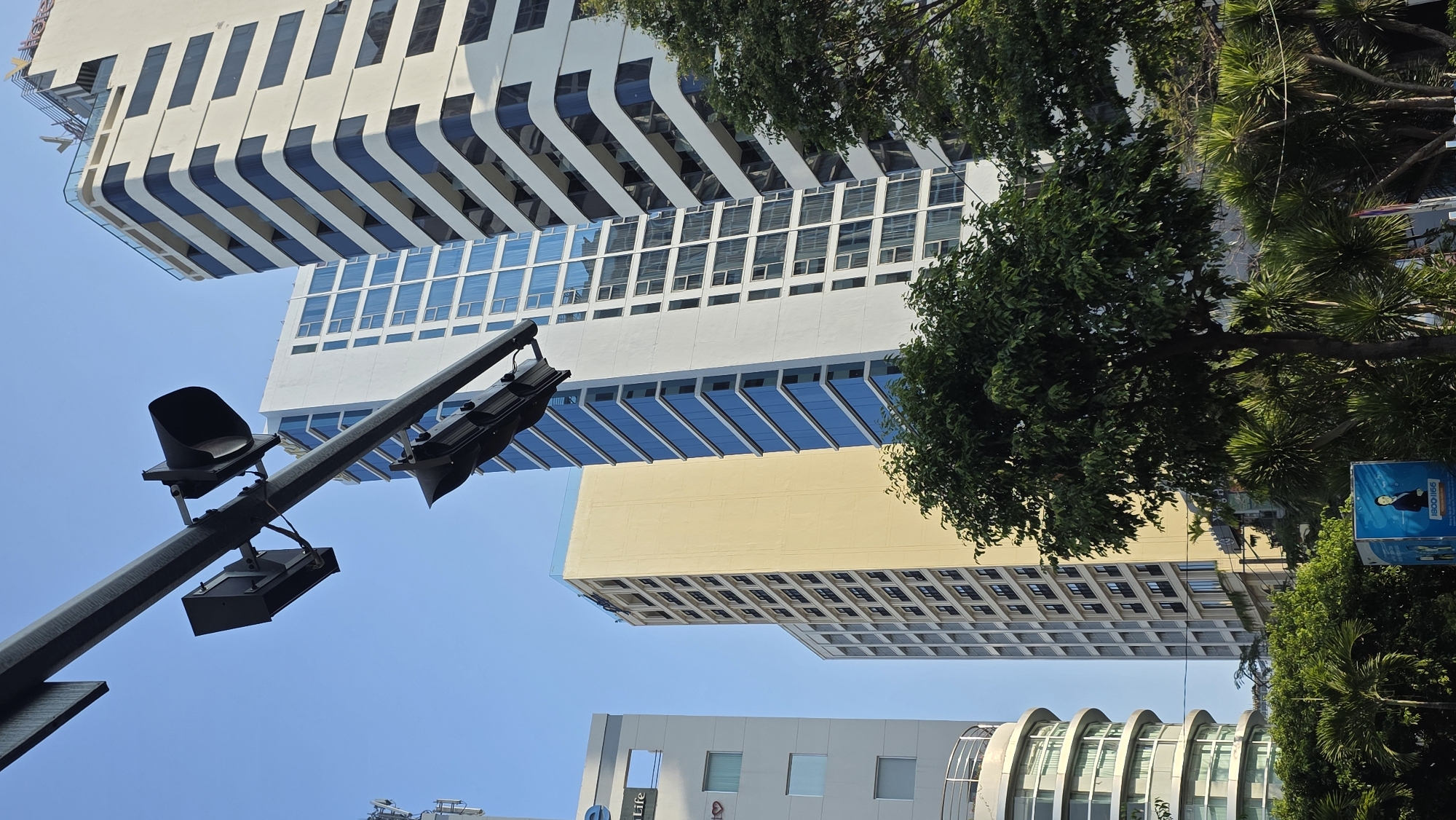 The image shows a low-angle view of buildings in Nha Trang, Vietnam. The buildings have a modern design, with multiple stories and glass windows. The sky is clear and blue. A black traffic light pole with lights is in the foreground. Trees and a sign are on the right side.