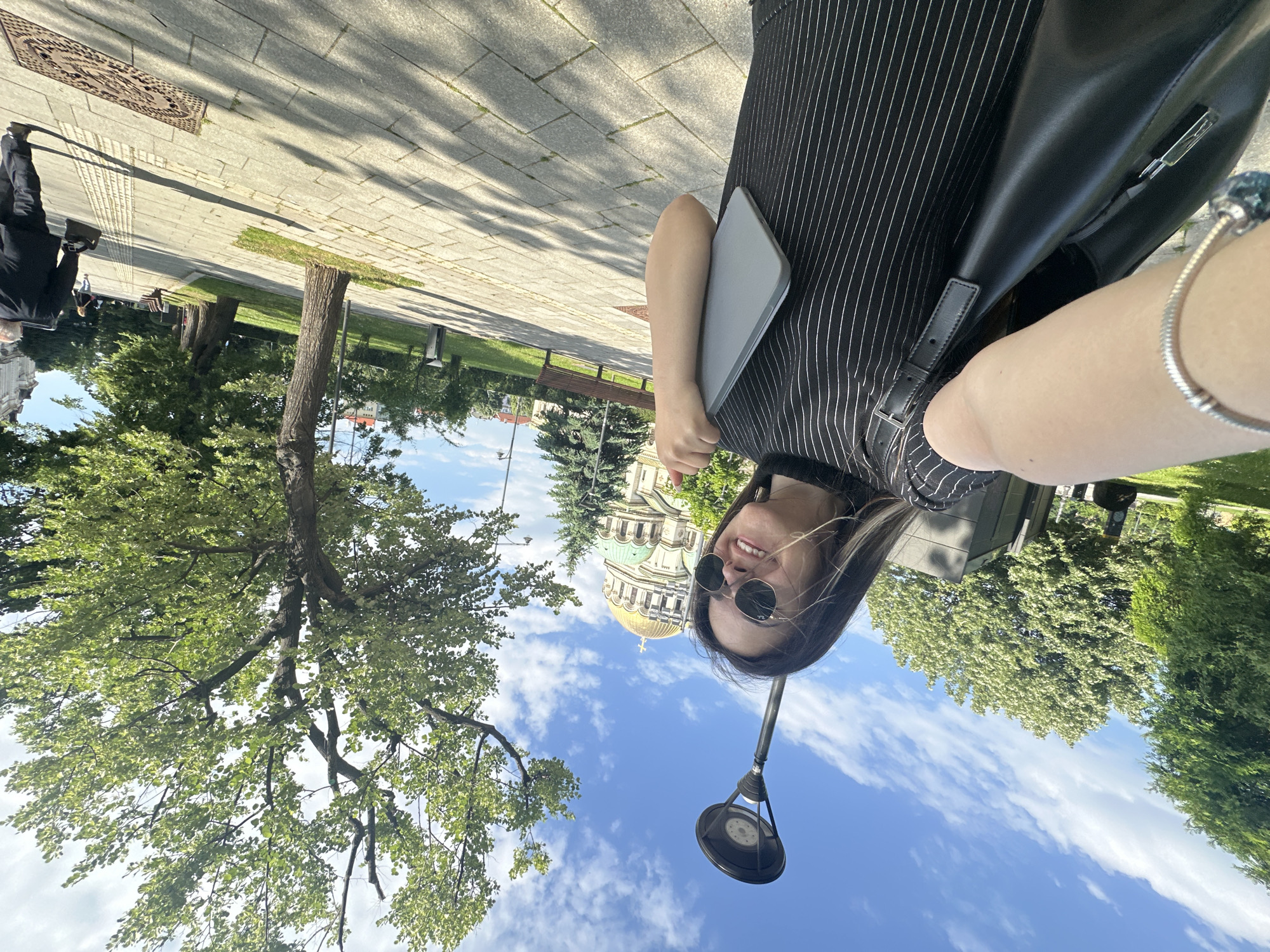 The image is a selfie of a woman wearing sunglasses and a striped black and white shirt. She is holding a tablet or notebook. Behind her is a view of the Cathedral of Saint Sava in Sofia, Bulgaria. The sky is blue with scattered clouds. A tree and a lamppost are visible in the foreground. The setting appears to be a paved area or sidewalk next to a park or green space. Another person in dark clothing can be seen in the far distance to the left.