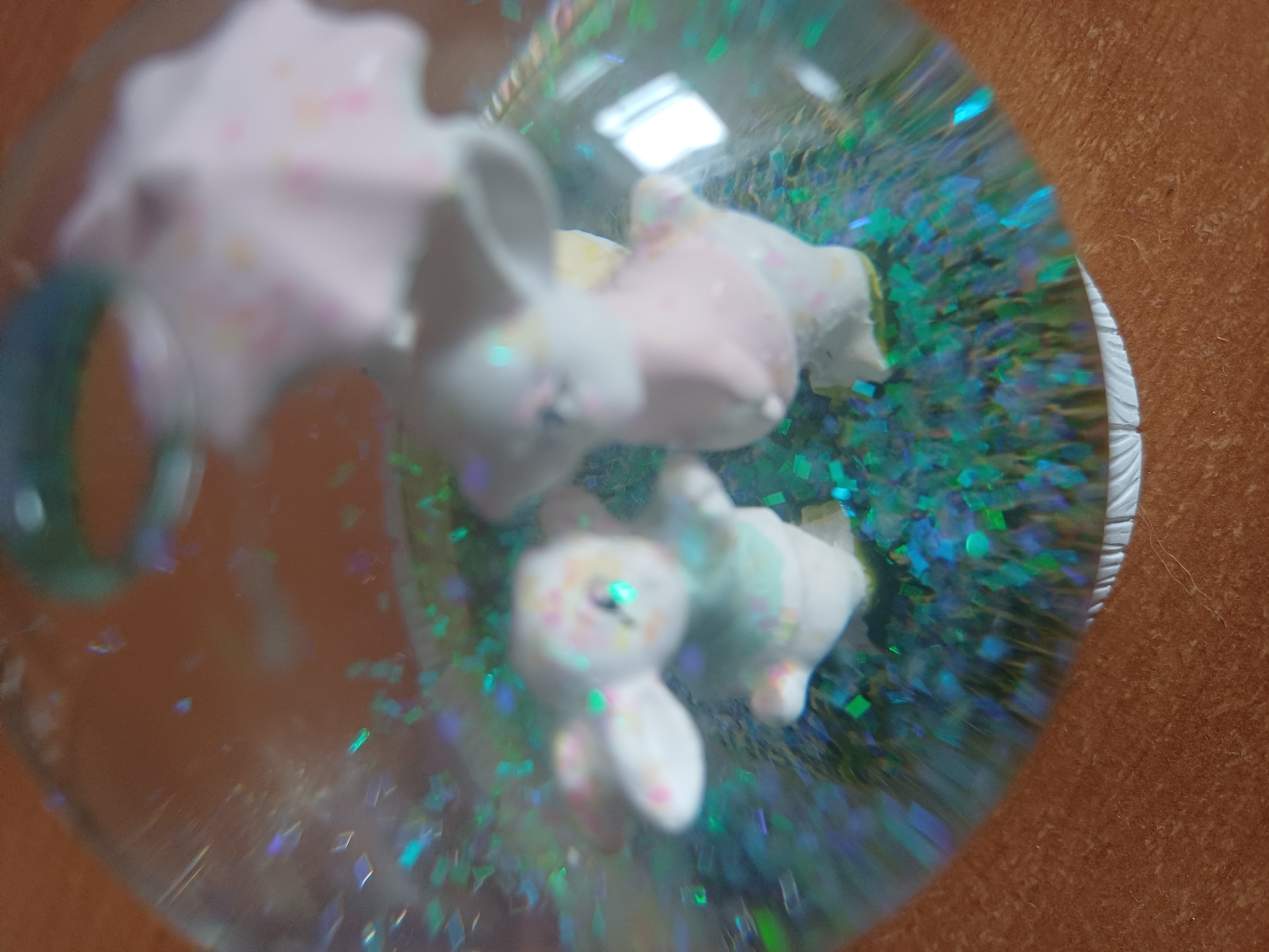 The image shows a close-up of a snow globe. Inside the globe, there are two small figurines of bunnies. One bunny is wearing a light blue outfit, and the other is wearing a pink outfit. Above the bunnies, there is a pink umbrella with small colorful dots. The interior of the snow globe is filled with clear liquid and numerous pieces of blue and green glitter. The snow globe is resting on a brown, textured surface, possibly a wooden table. The background is out of focus, but it appears to be a warm, indoor environment. There is no indication of the location being in Kyiv, Ukraine.