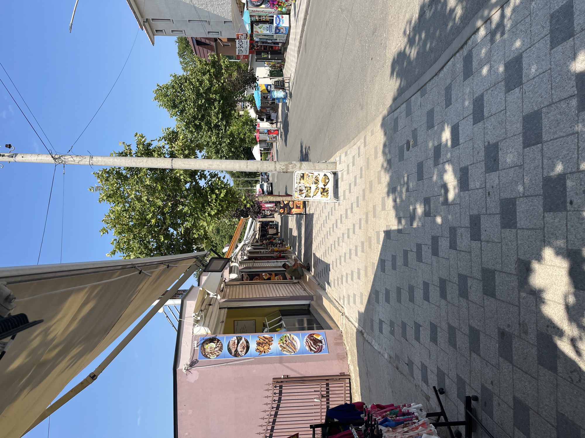 The image shows a street scene in Obzor, Bulgaria. On the left side of the street is a sidewalk with a pink building. The building has a metal fence and an awning that extends over the sidewalk. There are several shops along the sidewalk, including one with a sign displaying images of food. In front of this shop, there is a display rack with clothing. A person is sitting on the sidewalk in front of one of the shops.
On the right side of the street, there is a paved road with cars parked along it. There are more shops and kiosks on this side of the street, including one with a "TOP SHOP" sign. The shops have awnings and display various goods. A utility pole with wires runs along the sidewalk. Trees with green foliage are visible between the buildings and along the street. The sky is clear and blue, indicating a sunny day. Shadows of trees are cast on the sidewalk and the road.