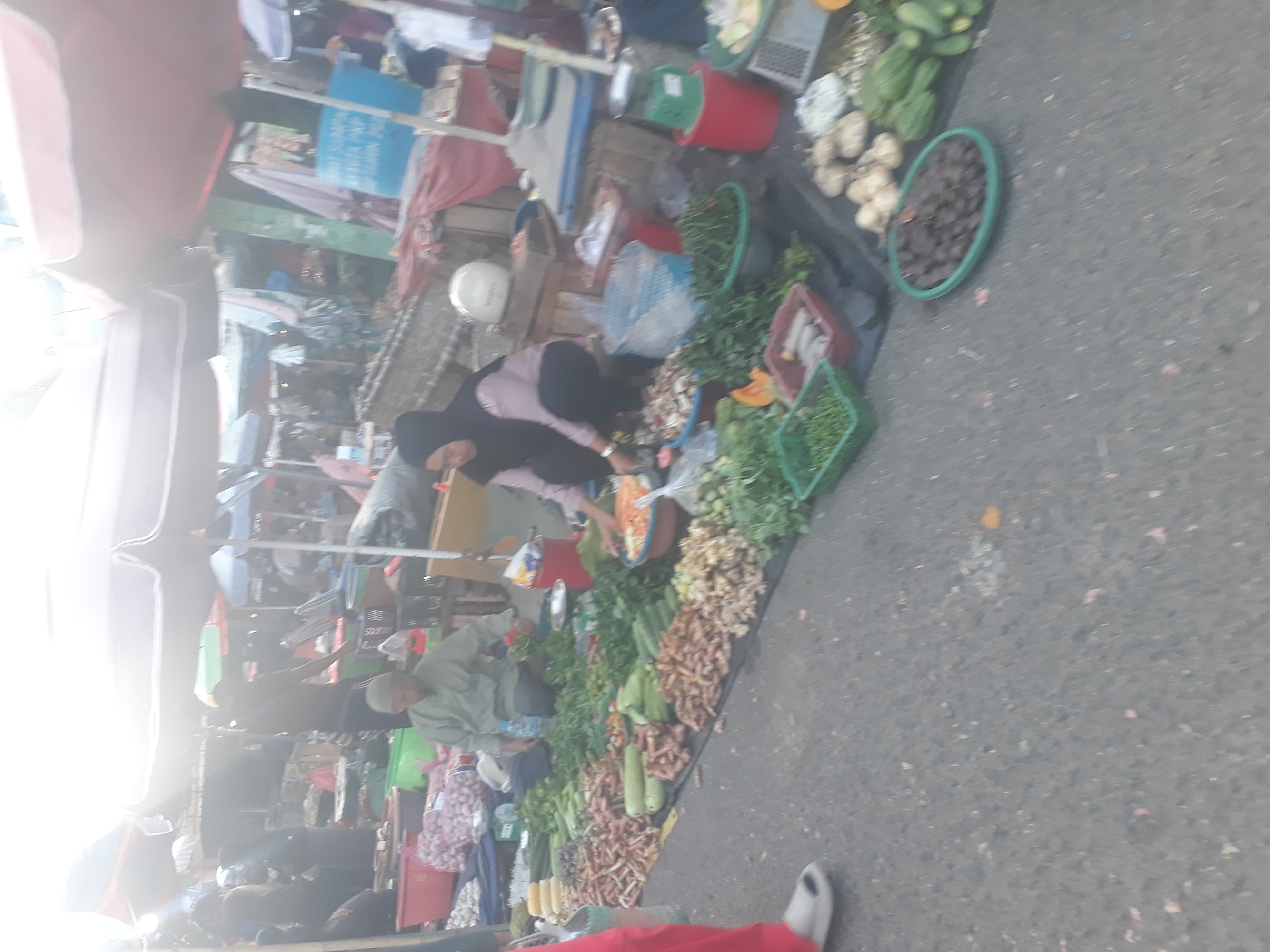 The image depicts an outdoor market scene in Pangkalpinang, Indonesia. In the foreground, a variety of fresh produce is displayed on a black tarp, including what appears to be ginger, turmeric, cucumbers, and leafy green vegetables. Several plastic baskets and bowls are used to hold some of the items. Two individuals are present: a man in a light-colored traditional garment and a white cap is crouched and interacting with the produce, and a woman in a black hijab and a pink shirt is also crouched, seemingly arranging or selling items. Behind them, more market stalls and awnings are visible, suggesting a bustling commercial area. The ground is paved asphalt, and debris is scattered across it.