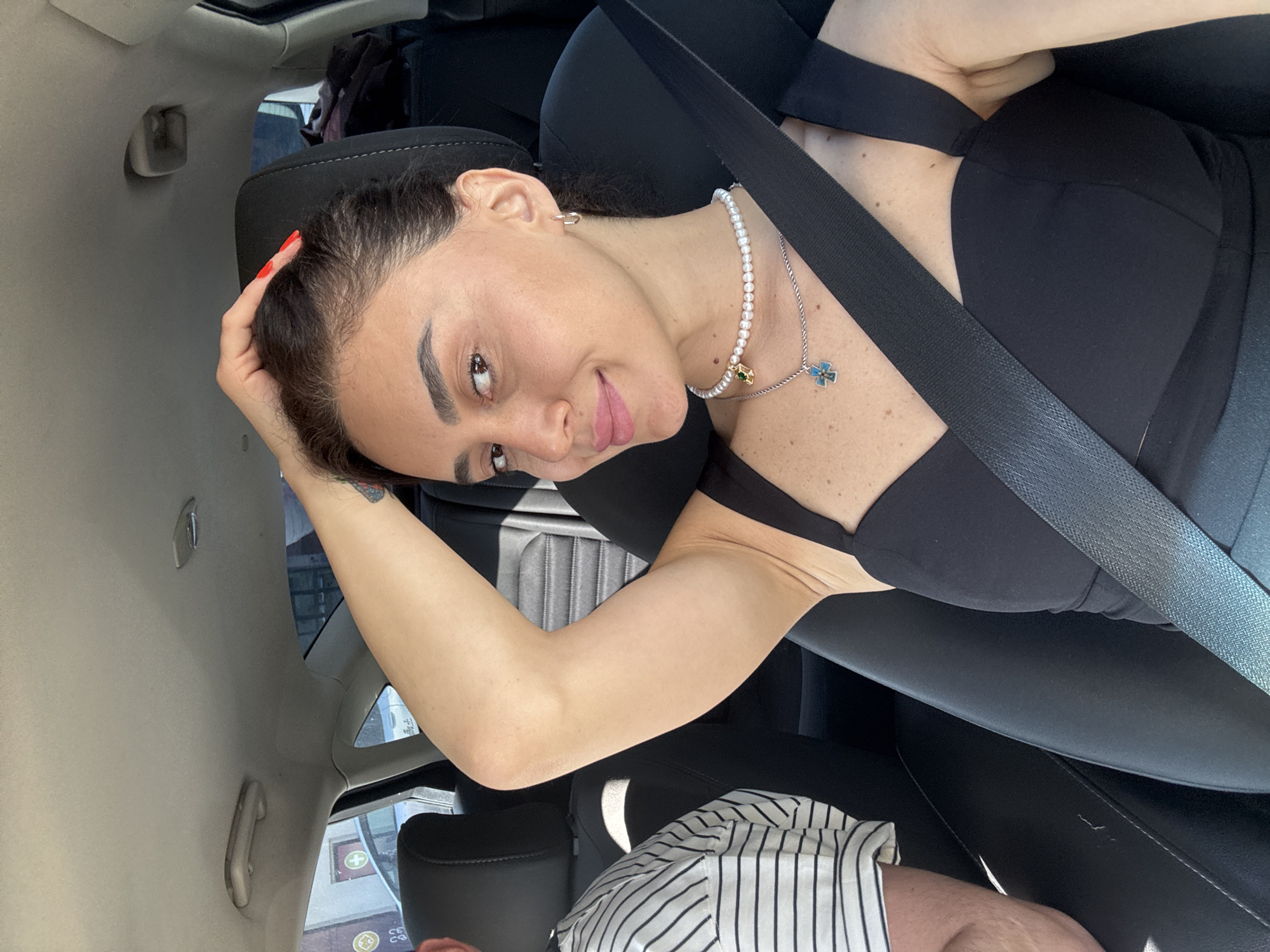 A woman with dark hair is sitting in the passenger seat of a car, her right arm raised and hand resting on her head. She is wearing a black dress and a pearl necklace with a pendant. A seatbelt is visible across her chest. The interior of the car is visible, including the headrest and part of the driver's seat, where a person in a striped shirt is seated. The car appears to be in motion, with a blurred background suggesting a city street. The sun is shining, casting shadows inside the vehicle.