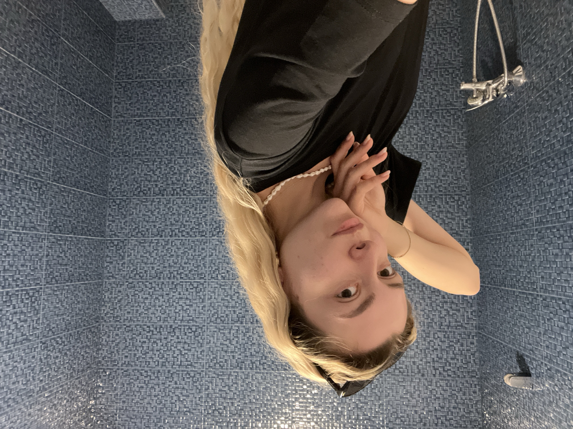 This close-up image features a young woman posed in what appears to be a tiled bathroom or shower. The original photograph is rotated 90 degrees clockwise, presenting the woman on her side with her head facing downwards.
When viewed upright, the woman, captured from the chest up, is looking directly at the camera with a calm, neutral expression. She has long, straight blonde hair that flows over her shoulders and fair skin. She is wearing a simple black top or dress, and a delicate white beaded necklace adorns her neck. A thin, golden bracelet is visible on her right wrist, and a pair of black sunglasses or a headband is pushed back on her head, partially visible against her hair. Her hands are gently brought together near her chest or chin, contributing to a soft and posed demeanor.
The background is uniformly covered with small, square mosaic tiles in a dark blue or gray-blue color, creating a textured surface. On the right side of the frame, a chrome-finished shower fixture, including a showerhead and controls, is mounted on the tiled wall. The bright lighting illuminates the scene, creating reflections on the glossy surface of the tiles. No visible text is present in the image.