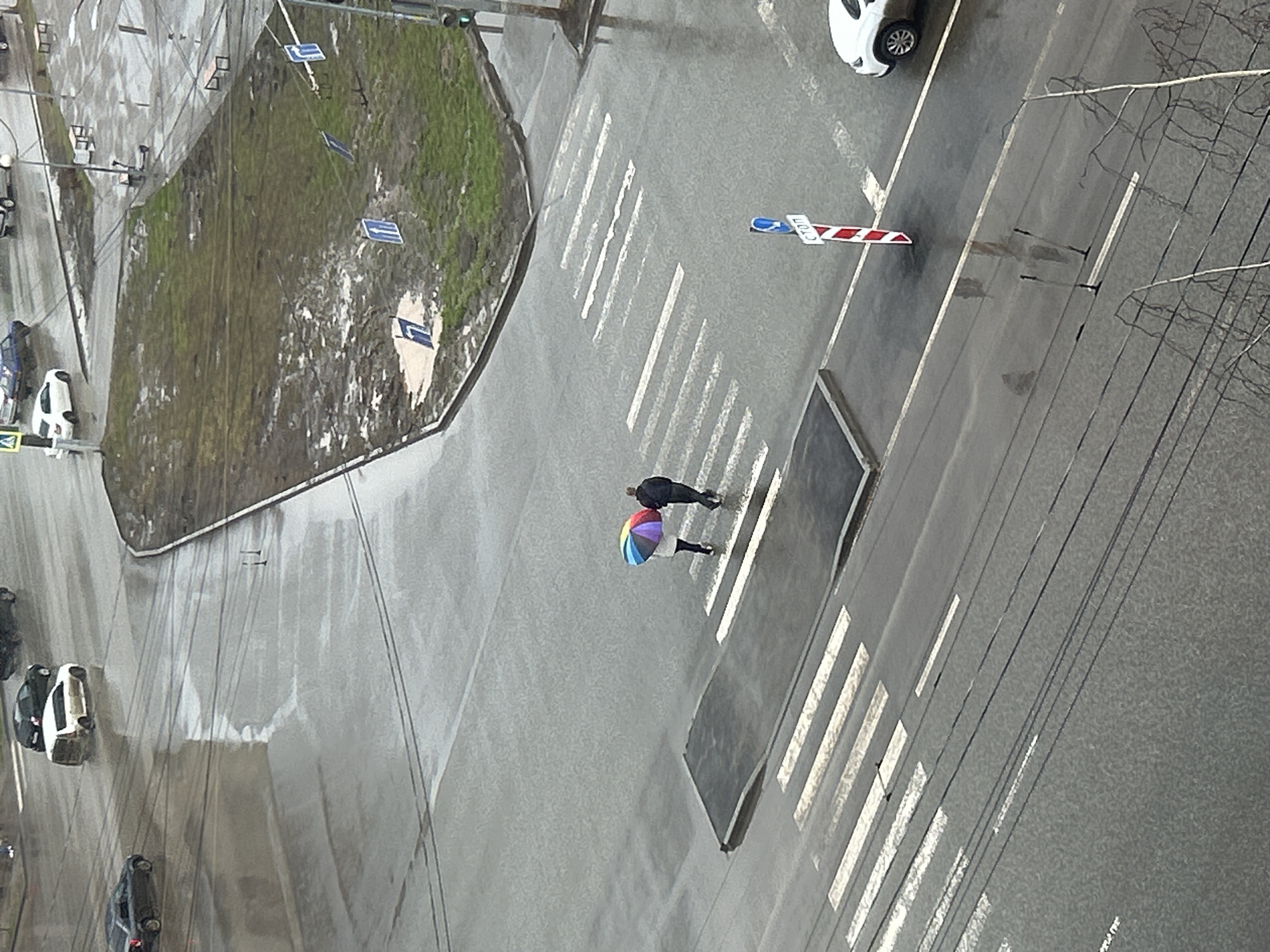 The image shows an aerial view of a street scene in Izhevsk, Russia on a rainy day. Two people are crossing a pedestrian crosswalk, one holding a colorful umbrella. Several cars are visible parked along the street and one is driving. A grassy area bordered by pavement lies adjacent to the crosswalk. Traffic lights and power lines are also visible. The scene appears wet due to the rain.