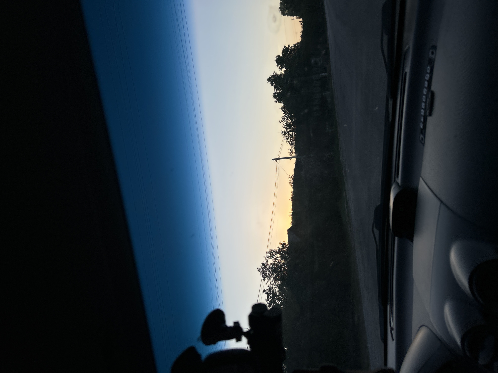 This image captures a view from inside a car, looking out through the windshield at a rural road during twilight. The sky is a gradient of deep blue at the top, transitioning to pale blue and then a soft yellow-orange near the horizon, indicating either sunrise or sunset. Power lines are visible crisscrossing the sky.
On either side of the road, there is dense foliage and trees, silhouetted against the sky. Several houses or structures are partially visible behind the trees, suggesting a village or small settlement. A power pole stands to the left of the road.
The foreground of the image shows the interior of the car. The dashboard is visible, with a dark, possibly plastic surface. A dashcam is mounted on the windshield in the upper left portion of the frame, its silhouette a prominent dark shape. The overall lighting suggests low light conditions. The scene appears to be in the vicinity of Basarabeasca, Moldova, given the rural setting and typical rural architecture. There are no people visible in the image.