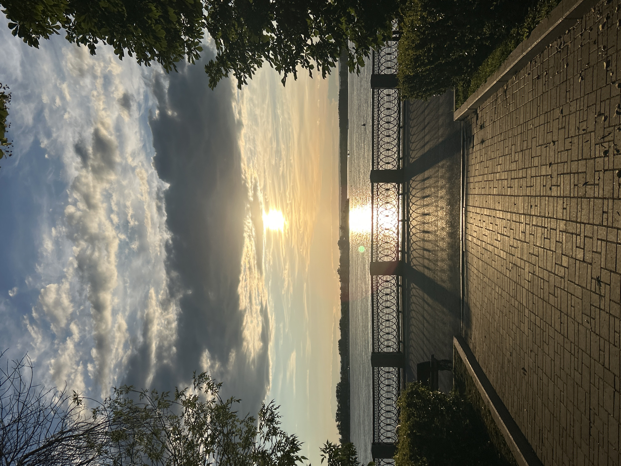 This media file depicts a scenic view of a waterfront promenade at sunset. The scene is captured in portrait orientation, with the sky and water dominating the left side of the frame and a paved walkway and decorative railing on the right.
The sky is filled with dramatic clouds, some dark and heavy, others illuminated by the setting sun, casting a warm, golden glow across the horizon. The sun itself is low in the sky, its rays reflecting brilliantly on the surface of a calm body of water, likely a river or lake. The water appears placid, with subtle ripples catching the light.
A black metal railing with an ornate circular pattern separates the walkway from the water. The railing casts long, sharp shadows across the tiled pathway, indicating the low angle of the sun. The pathway is paved with rectangular bricks laid in a geometric pattern, giving it a structured appearance. Green foliage from trees frames the top and bottom edges of the image, adding a natural element to the urban setting.
The time of day is clearly sunset, with the warm, golden light and long shadows being prominent indicators. The weather appears to be clear with scattered clouds, creating a visually appealing sky. The presence of a promenade, railing, and paved walkway suggests an urban or semi-urban waterfront area. No people are visible in the frame. The overall mood is peaceful and serene, with the natural beauty of the sunset creating a tranquil atmosphere.
There is no visible text in the image. The location context is Novokuybyshevsk, Russia.