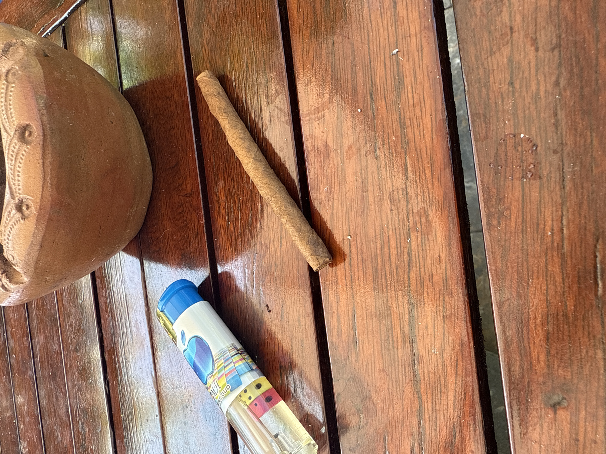 The image shows a wooden table with a cigar and a lighter. The table has a glossy finish and appears to be outdoors, possibly on a patio or deck. In the background, a part of a terracotta pot is visible. The lighting suggests it is daytime. The overall scene is simple and uncluttered, focusing on the two main objects on the table. The location is not explicitly stated to be in Cam Lâm, Vietnam; this information is only provided as context.