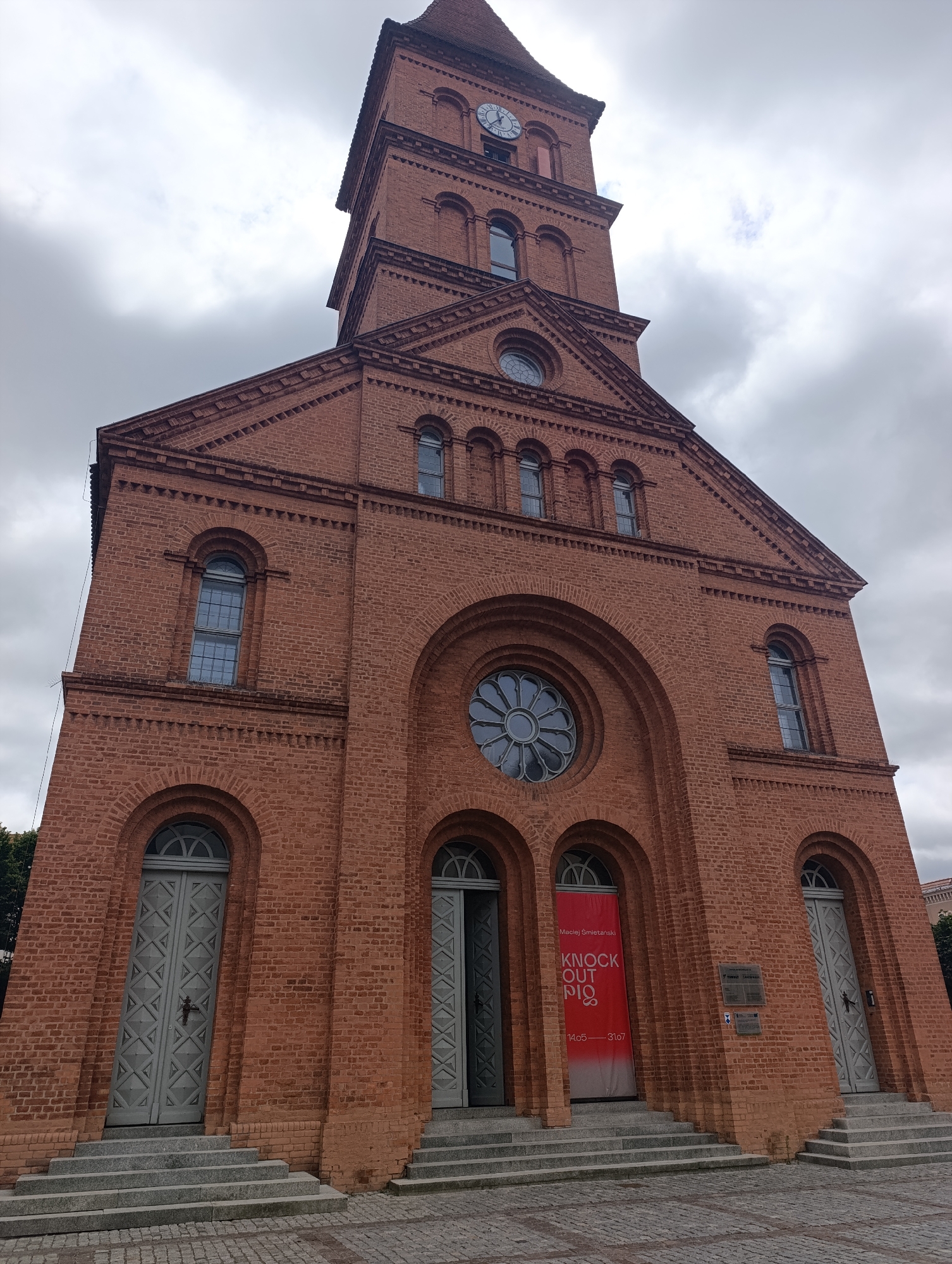 The image shows the exterior of a brick church, likely the Church of the Holy Spirit in Toruń, Poland. The church has a tall tower with a clock face. The facade features several arched windows and doors. A red banner with white text is hanging on the left side of the entrance. The sky is overcast with clouds. The ground in front of the church is paved with cobblestones.