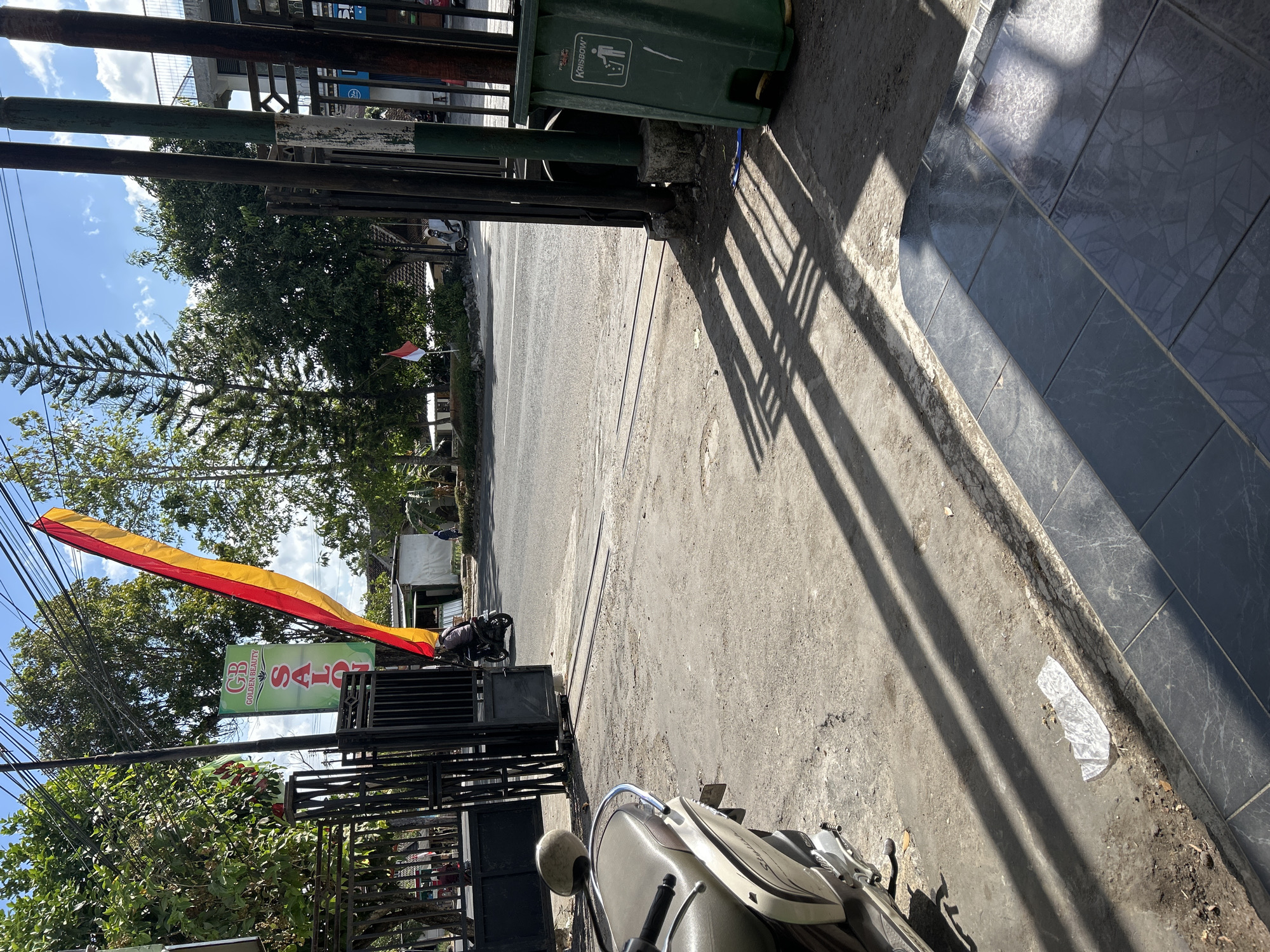 The image shows a street scene in Wonosari, Indonesia, on a sunny day. In the foreground, a scooter is parked on a paved area. A tall, green trash can is visible to the right, with a "Krisbow" logo. A red and yellow flag hangs prominently from a pole, likely associated with a business. A sign for "GOLDEN BEAUTY SALON" is visible behind the flag. A motorcycle with a rider passes through an open gate on the left side of the frame. The street is paved and appears to be a public road with some traffic. Trees line the sides of the street, and a building with a railing is visible in the background. The overall impression is of a typical street view in a town or city.
