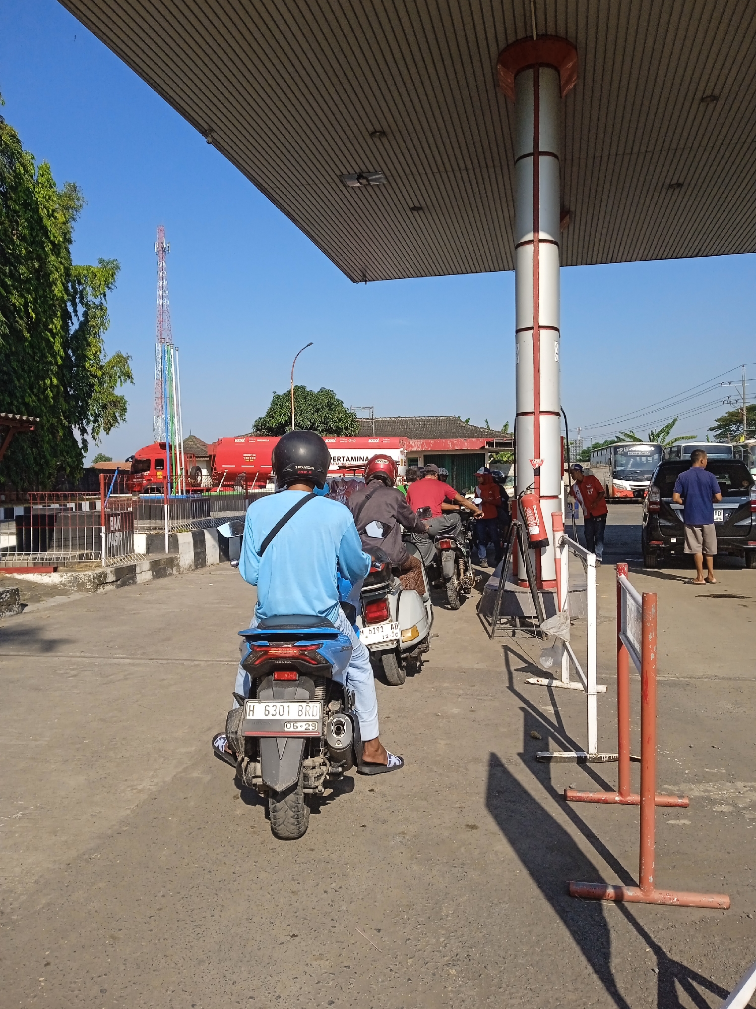 The image depicts a line of motorcycles at a Pertamina gas station in Brangsong, Indonesia. The foreground shows the back of a blue motorcycle with a license plate reading "H 6301 BRD". The rider wears a blue shirt and a black helmet. Behind the blue motorcycle, other motorcycles are waiting. A red Pertamina truck is visible in the background, along with other vehicles and people filling up with gasoline. A white column supports the gas station's roof, which casts a shadow on the ground. The sky is clear and blue.